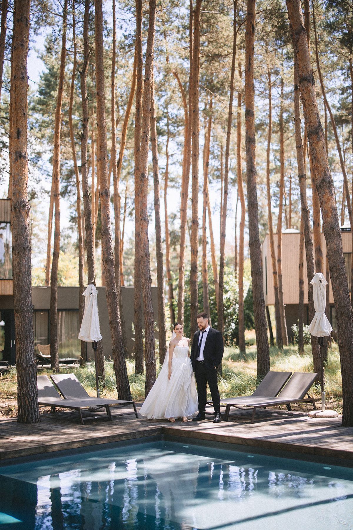 A couple stands beside a serene swimming pool surrounded by tall pine trees at an outdoor forest venue.