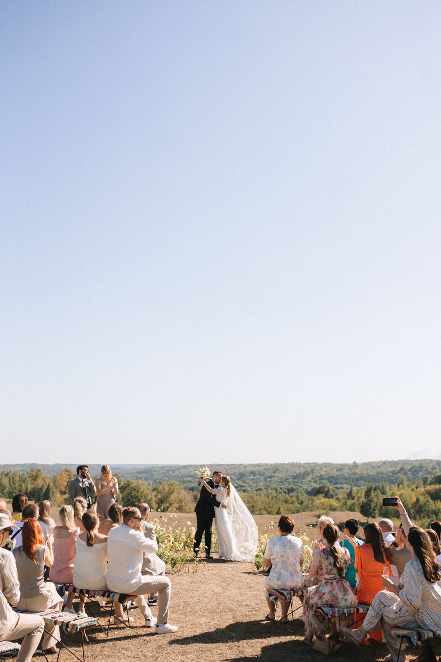 Outdoor hilltop wedding ceremony with guests seated on benches overlooking a scenic valley landscape on a sunny day.