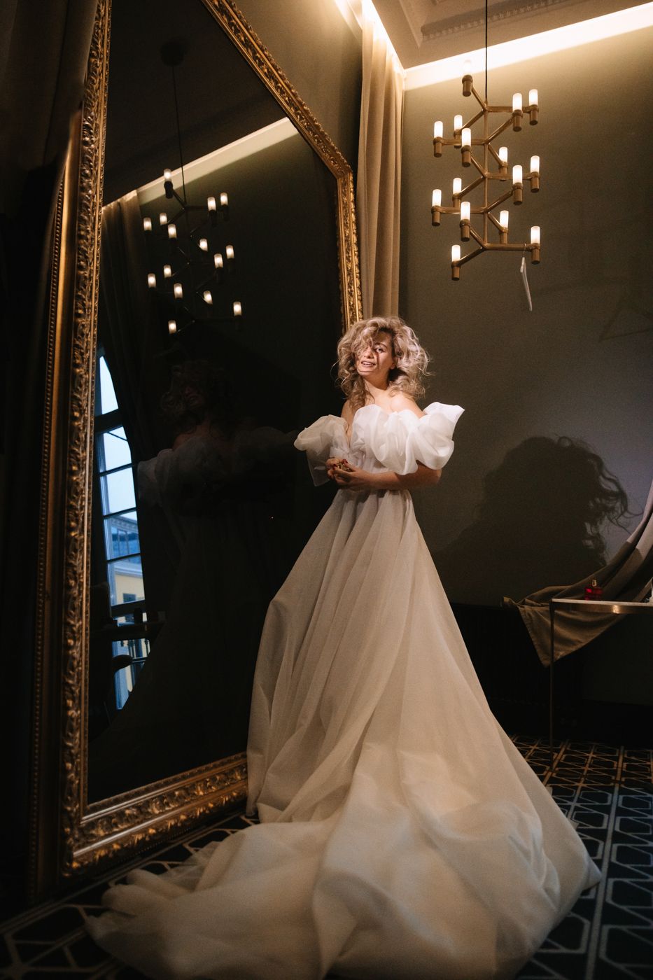 Wedding dress silhouette against ornate mirror and hanging lights in dark room.