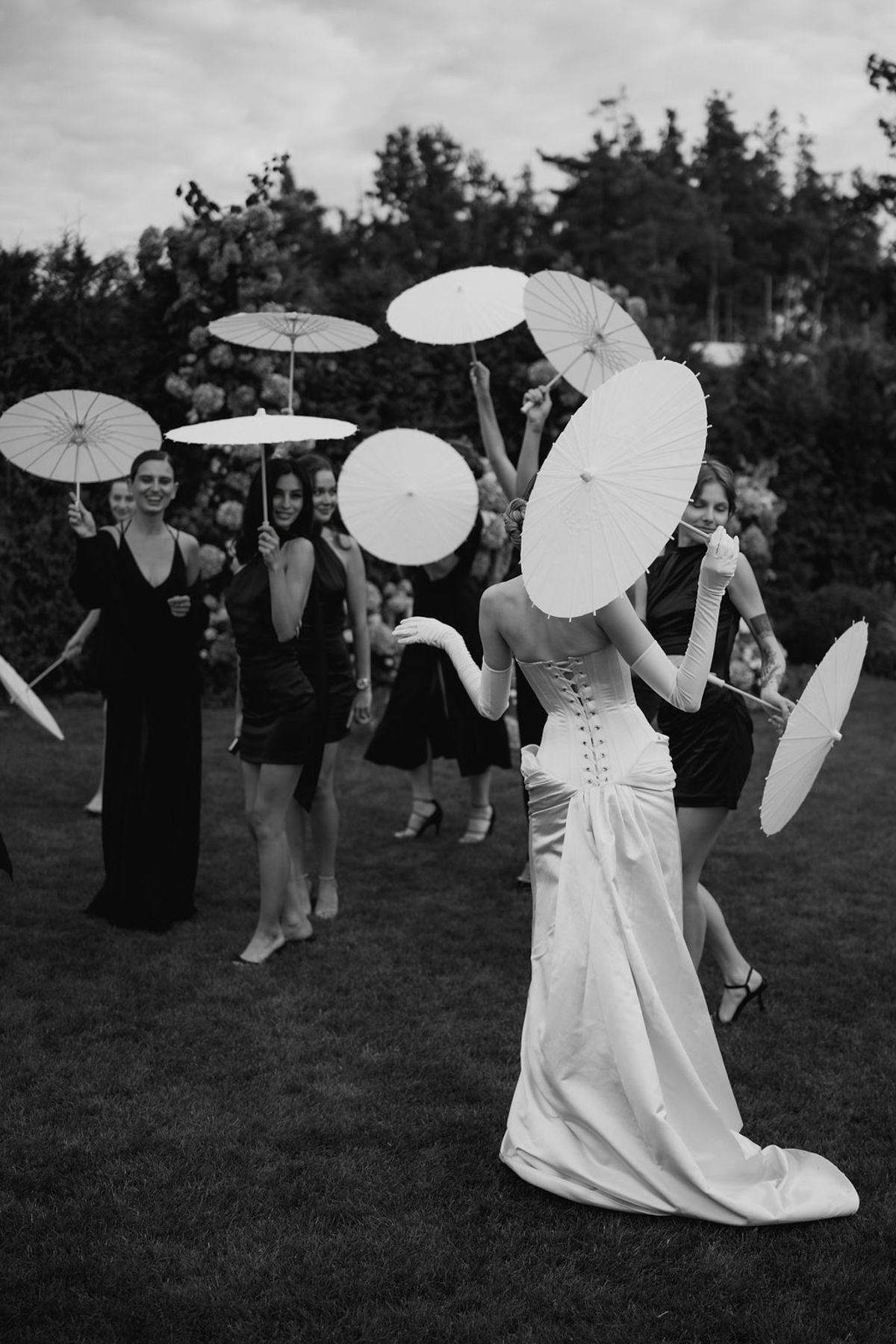 Wedding party members twirl white parasols while dancing on a grassy lawn in black and white photography.