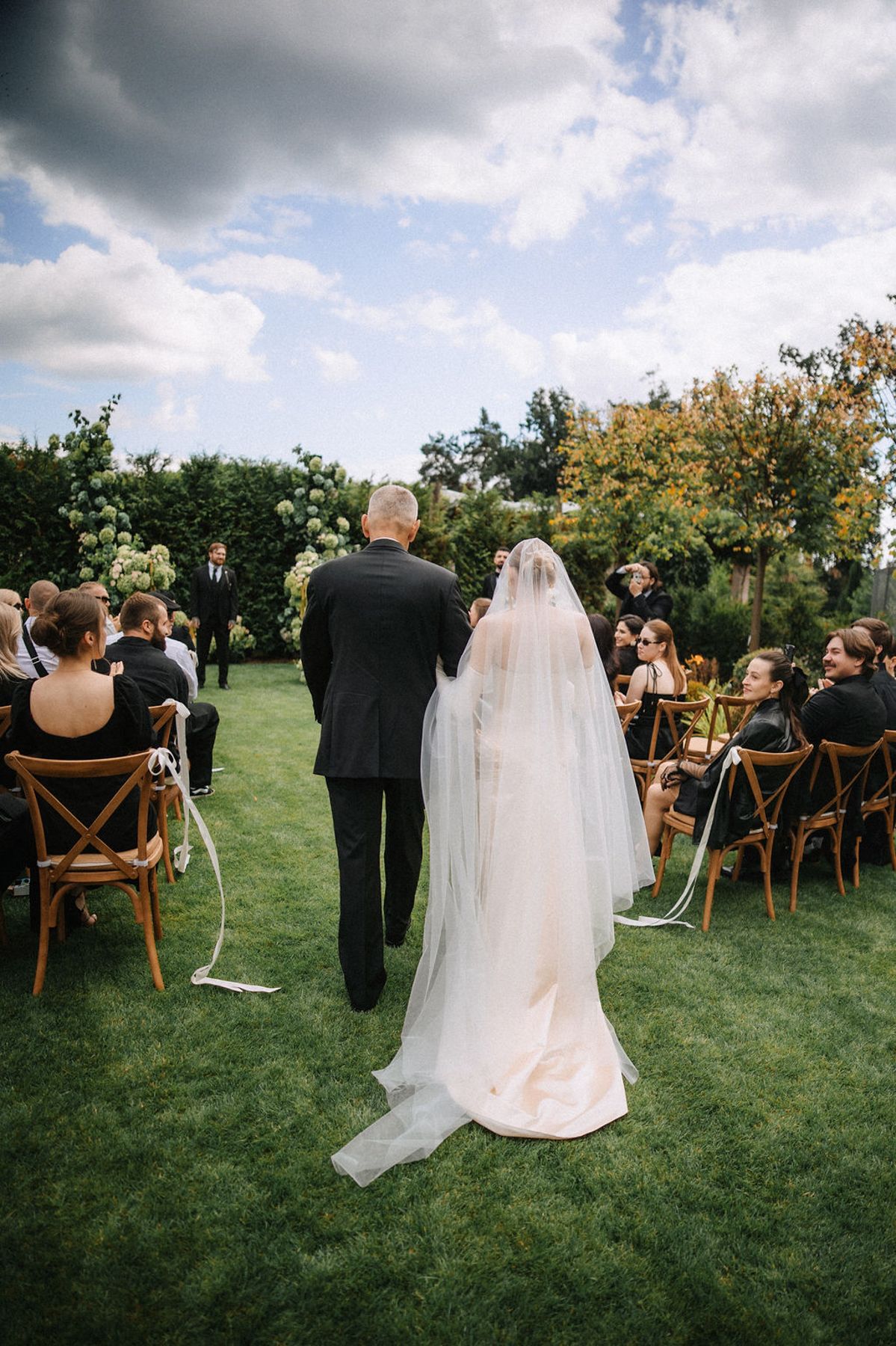 Outdoor wedding ceremony setup on a green lawn with wooden chairs under cloudy skies during a processional moment.