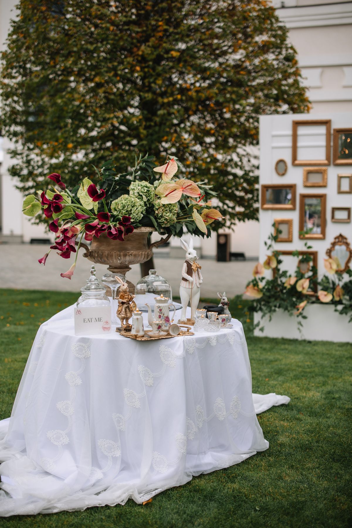 Elegant outdoor tea party setup with white tablecloth, floral centerpiece and vintage frames displayed on wall.