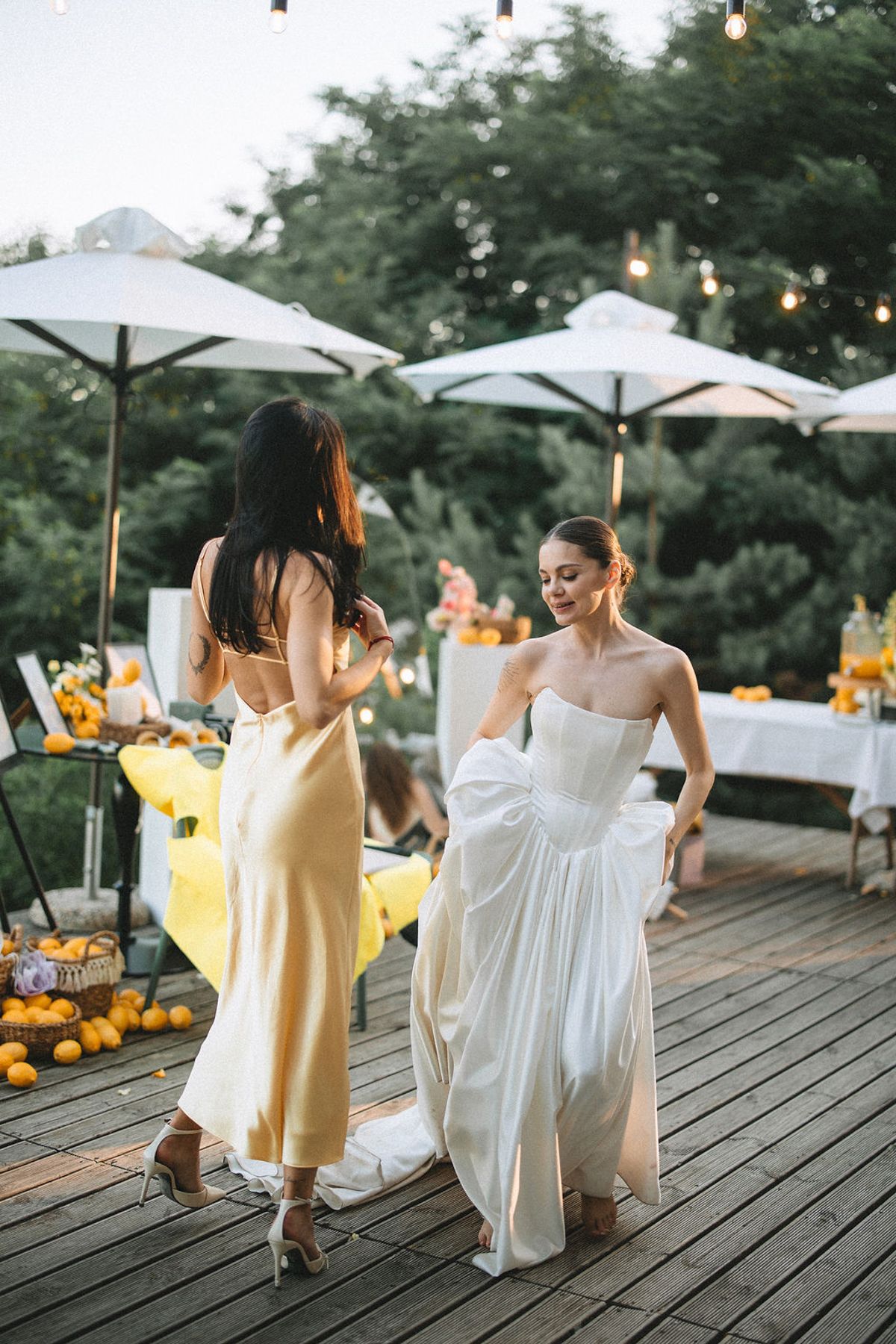Two women in elegant dresses dance on a wooden deck decorated with umbrellas and citrus fruits at an outdoor event.