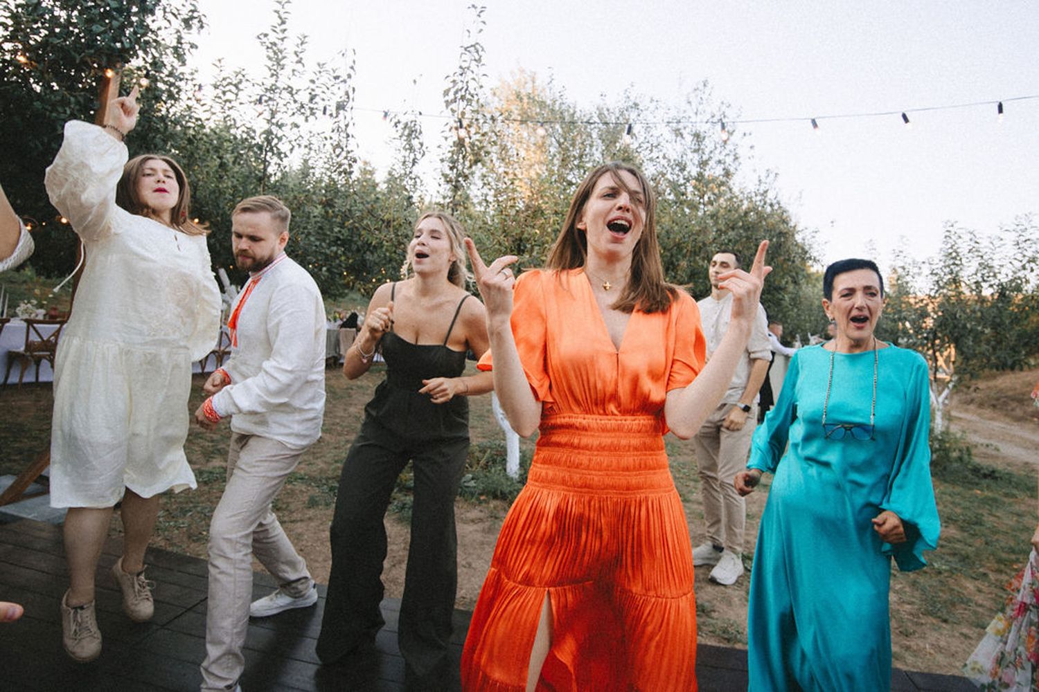 A group of wedding guests dance joyfully at an outdoor reception in colorful orange and teal dresses.