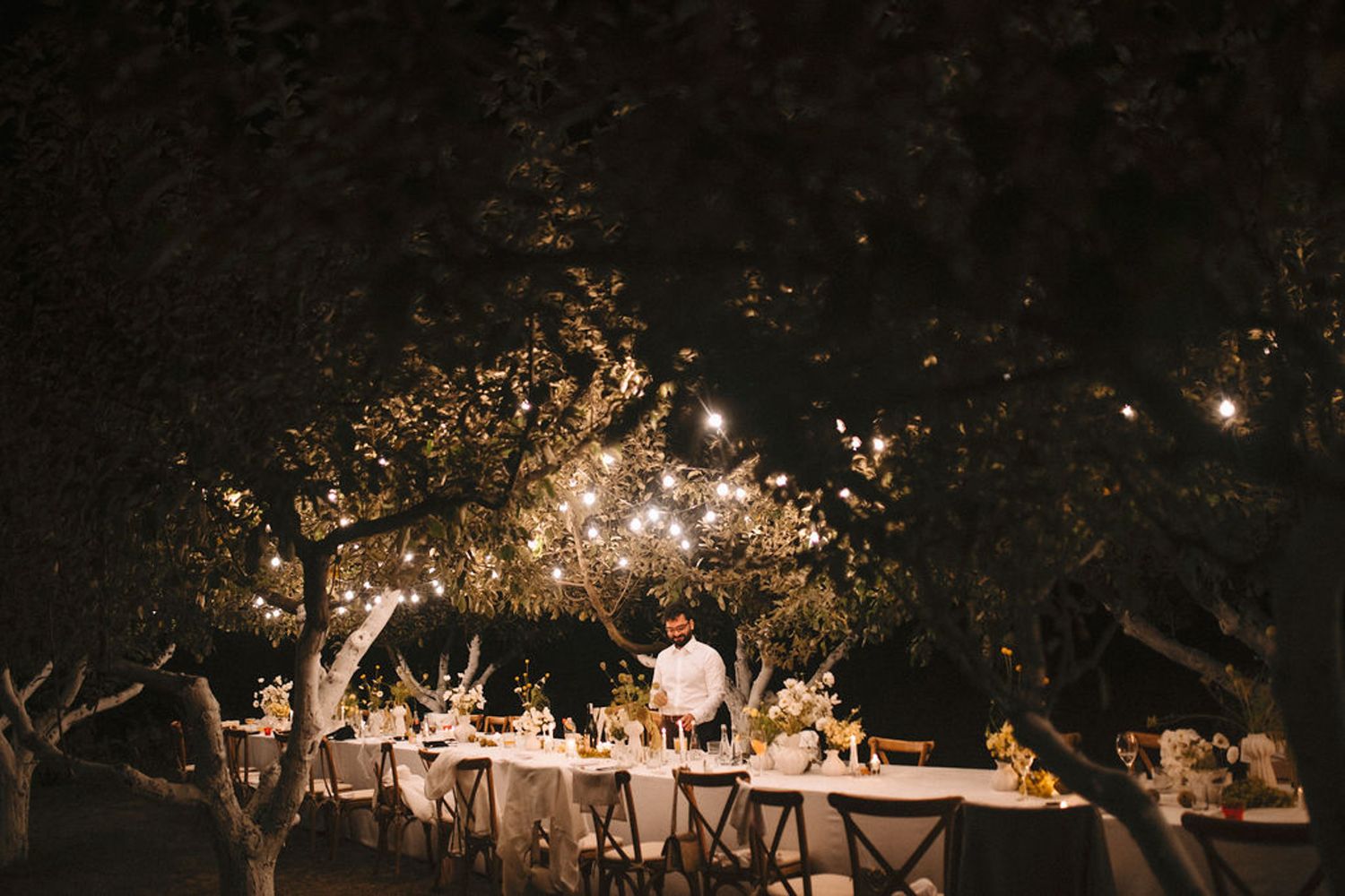 A romantic outdoor wedding reception setup with string lights illuminating tables under trees at night.