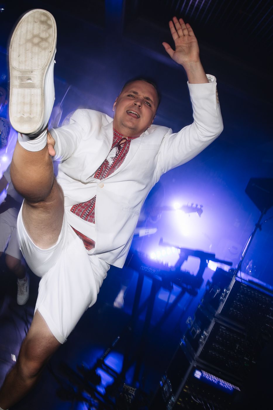 Energetic performer in white outfit with red tie dances on stage under dramatic blue lighting.