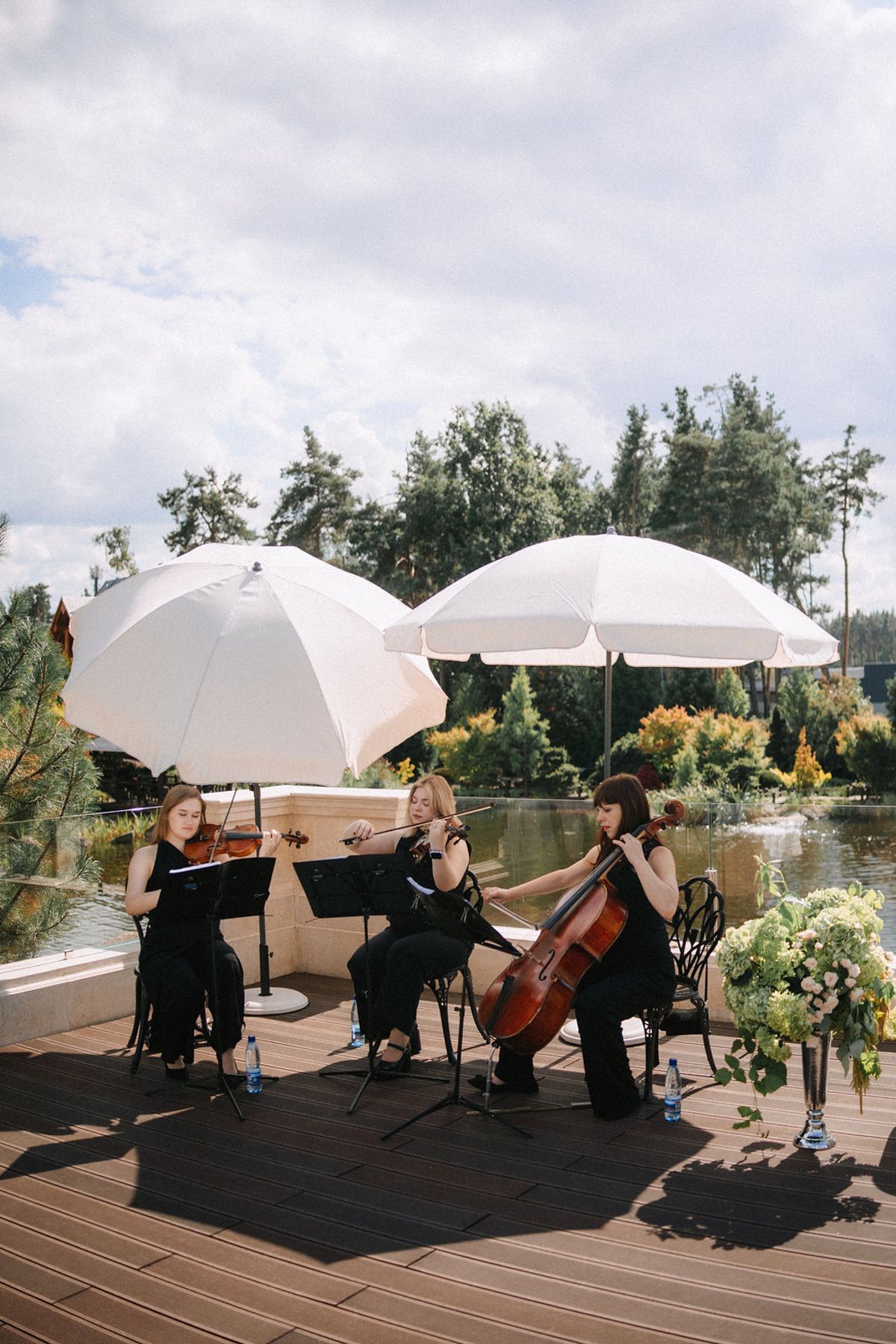 String quartet musicians perform outdoors under white umbrellas on a wooden deck by a lake with trees in background.