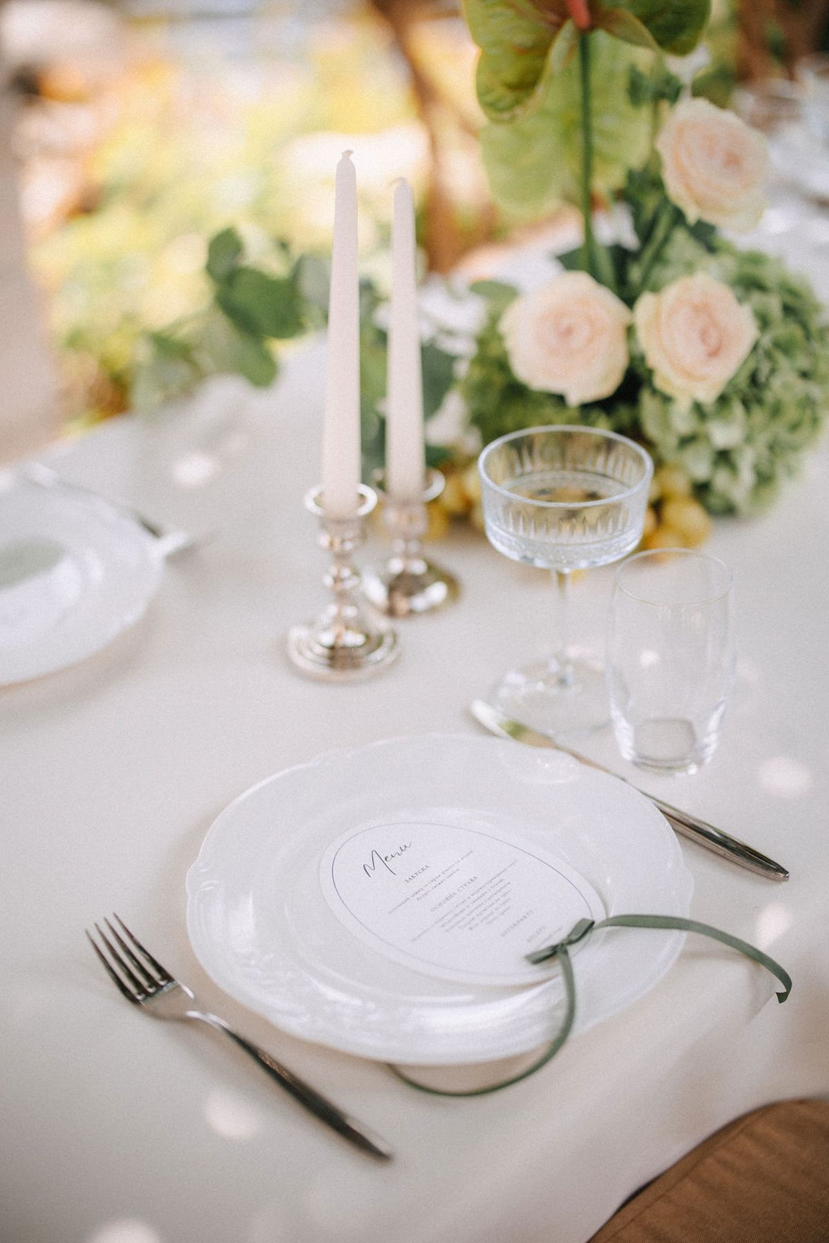 Elegant wedding table setting with white plates, silver candlesticks, pale pink roses and greenery on white tablecloth.