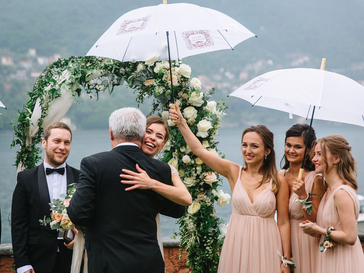 Wedding party stands together under white umbrellas during a rainy outdoor ceremony by the water.