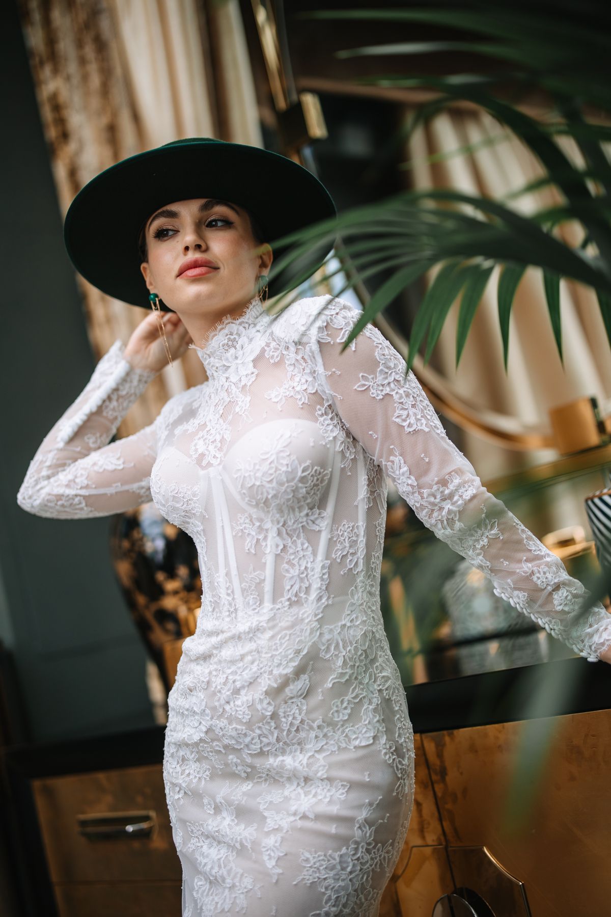 Elegant bridal fashion photo featuring a white lace dress and dramatic black hat in a modern interior setting.