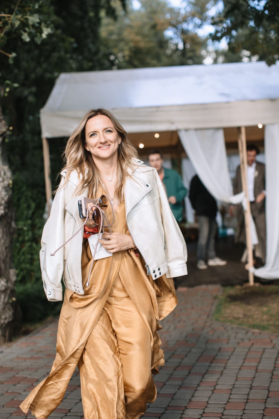 A person in a gold dress and white leather jacket dances joyfully at an outdoor event with white tents in the background.