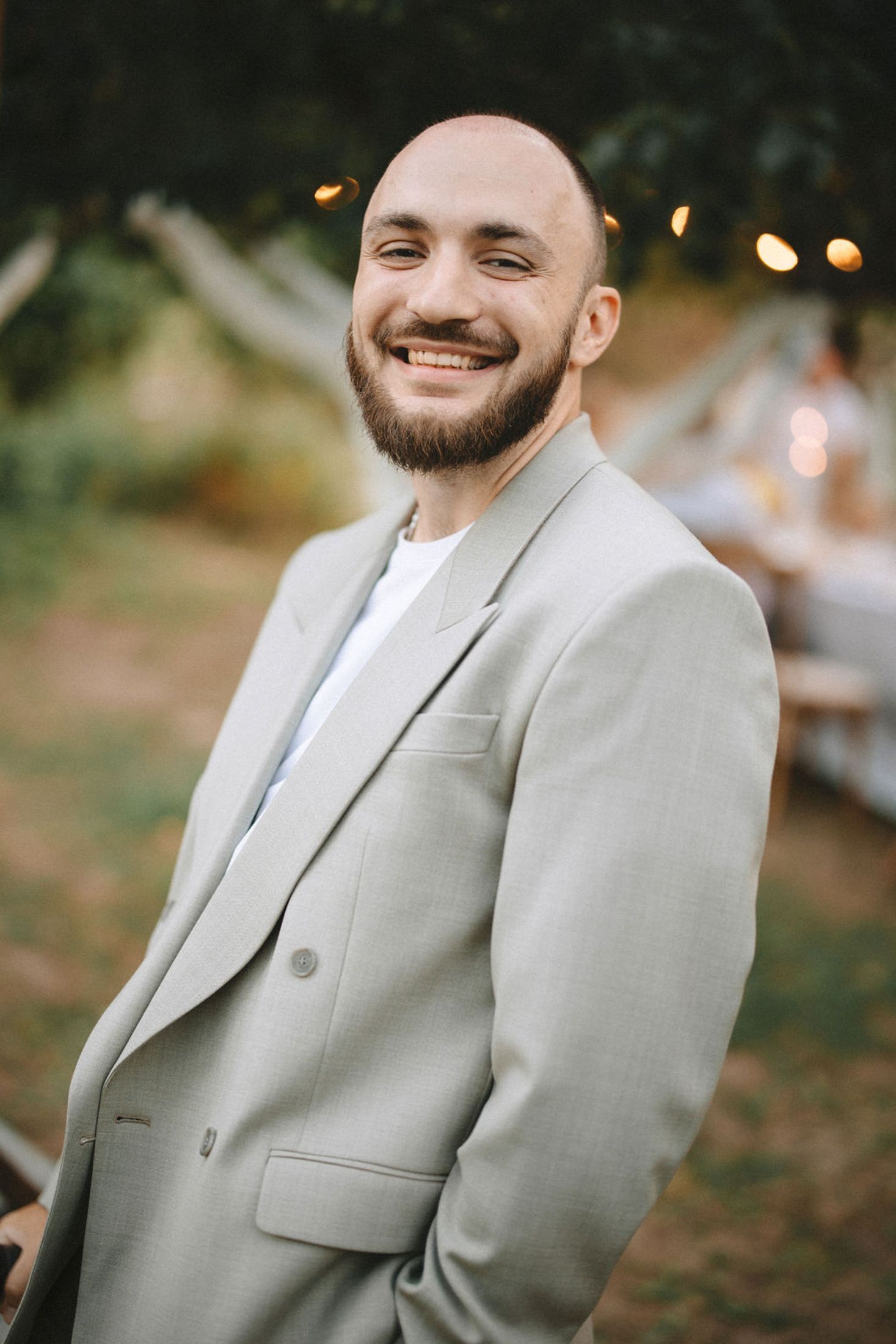 A person in a light gray suit jacket and white shirt smiles warmly at an outdoor event with twinkling lights in background.