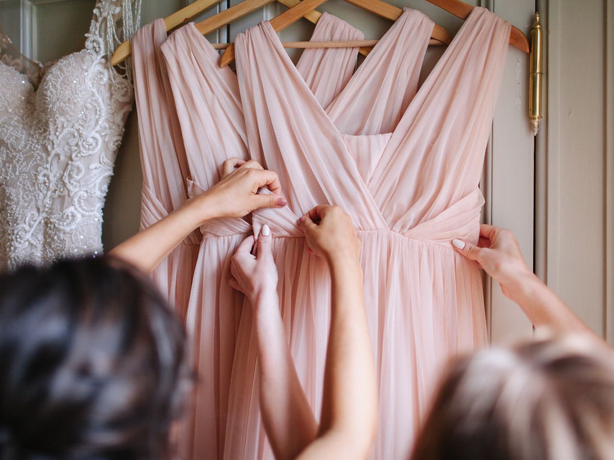 Blush pink bridesmaid dresses hanging on wooden hangers against a light colored wall.