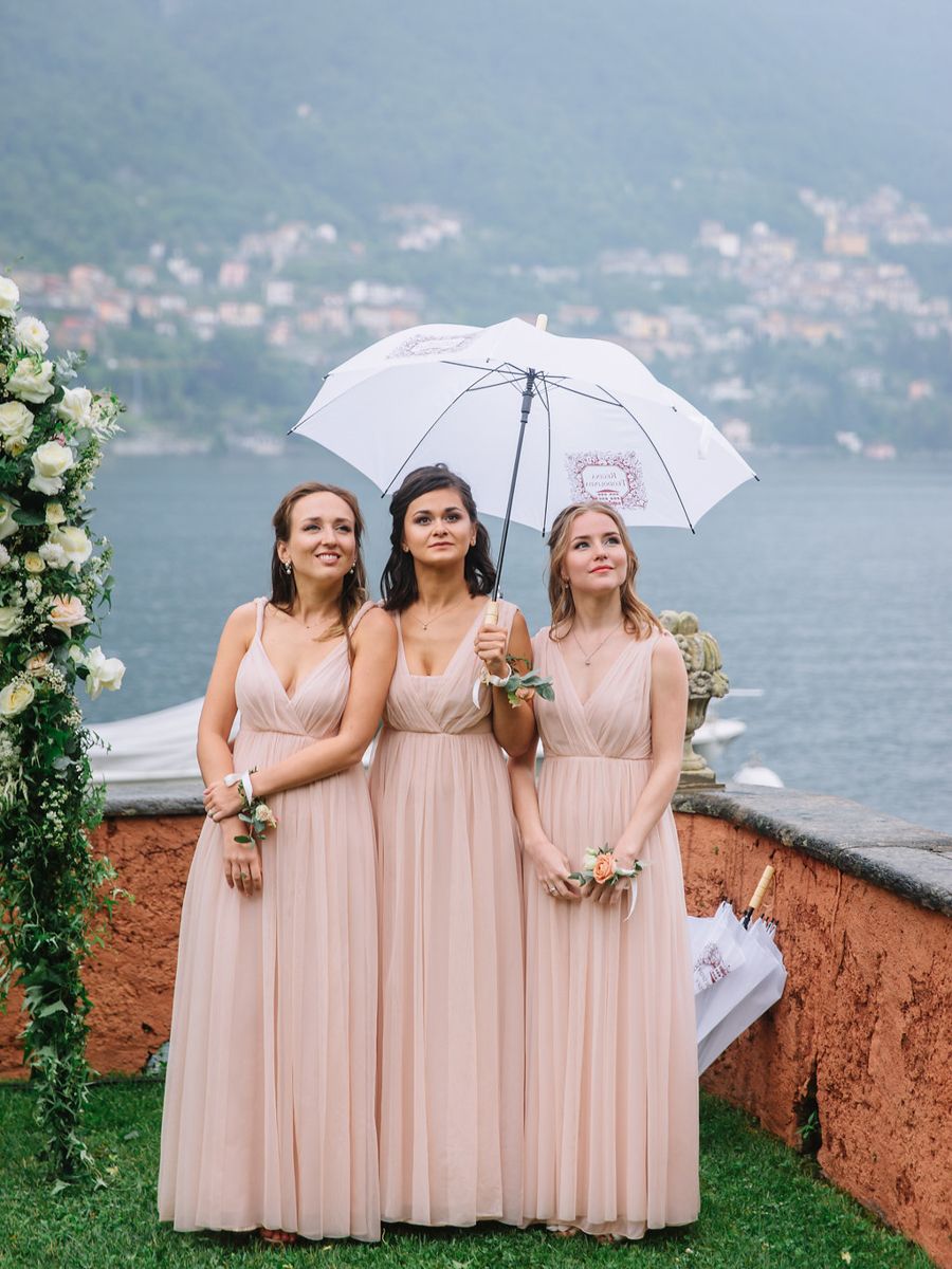 Three bridesmaids in blush pink dresses pose together on a terrace overlooking a scenic lake and hillside village.