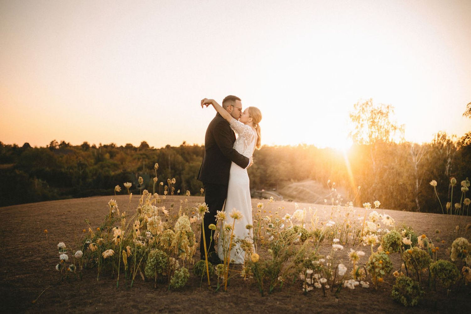 Couple in formal attire embracing at sunset in a field of wildflowers with golden sunlight streaming through the trees.