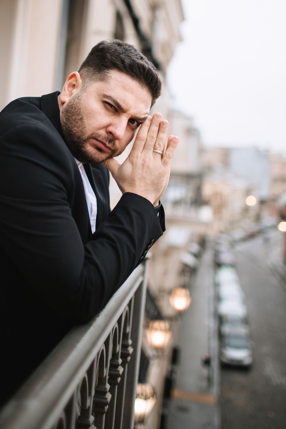 Person in dark suit leans thoughtfully against balcony railing in urban setting.