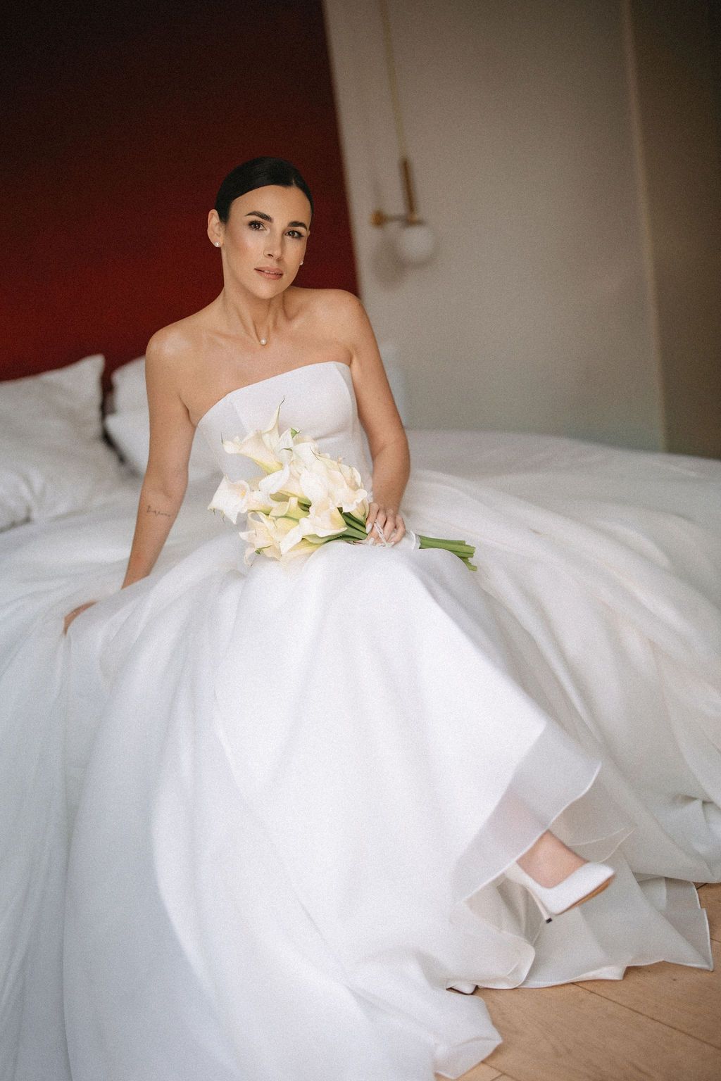 Bride in strapless white wedding gown sits gracefully on bed holding elegant white lily bouquet.