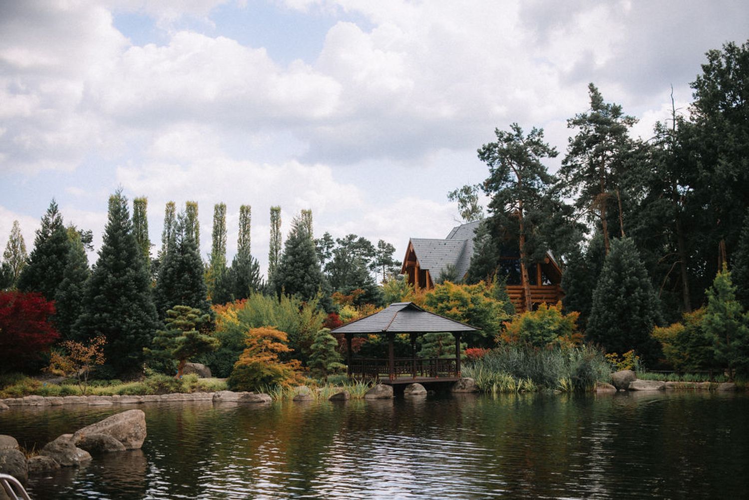 Tranquil Japanese garden with pagoda gazebo overlooking serene pond surrounded by evergreen trees and autumn foliage.