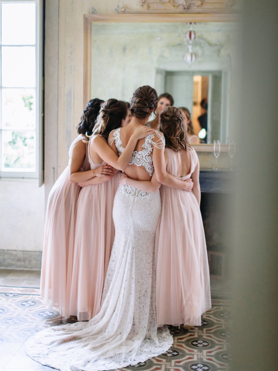 A bride in a lace gown hugs with bridesmaids wearing blush pink dresses in an elegant room with ornate details.