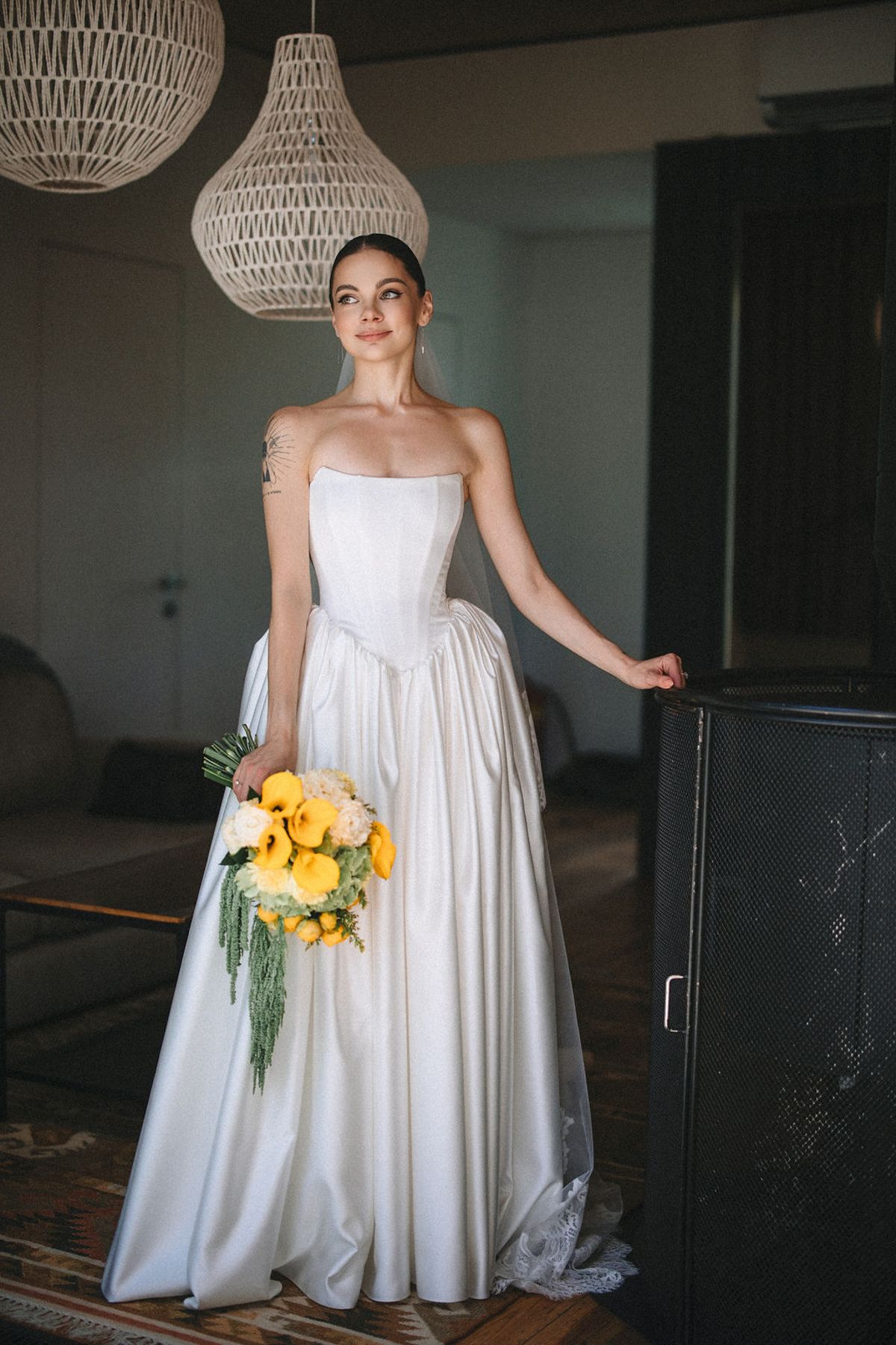 A bride in a white strapless gown holds yellow flowers while standing in a dimly lit room with hanging pendant lights.