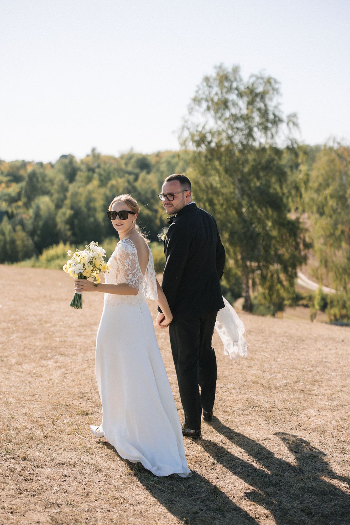 A couple in wedding attire stands on a hilltop during golden hour with trees in the background.