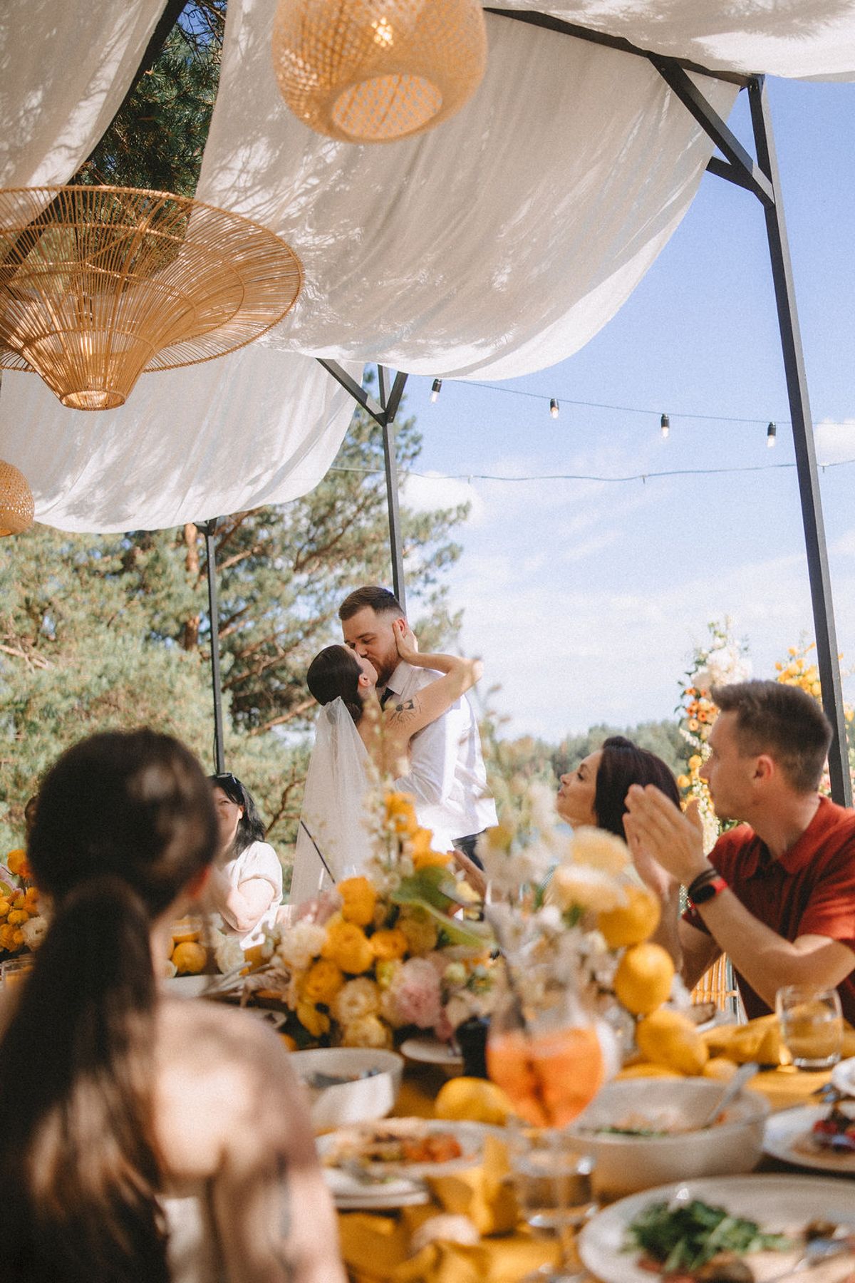 People gather at an outdoor dining table under white fabric canopies and woven pendant lights for a festive meal.