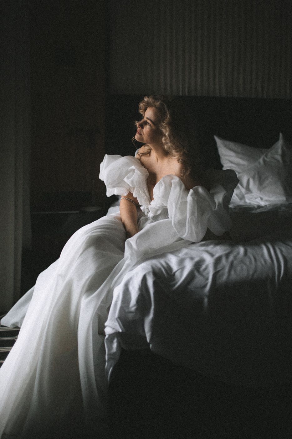 Dramatic boudoir photo in white dress against white bedding in dark room.