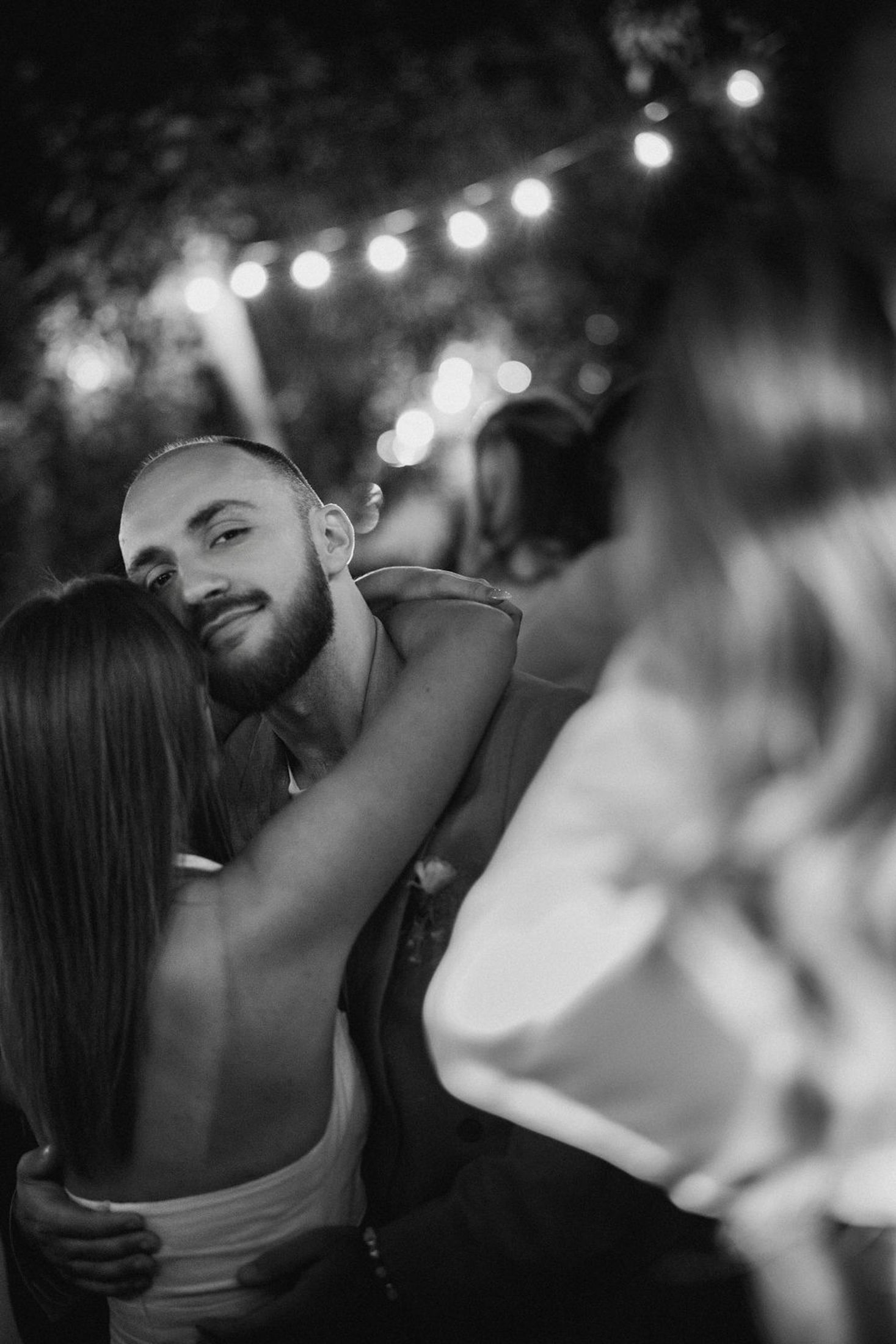 A romantic black and white moment at an evening celebration with string lights glowing in the background.