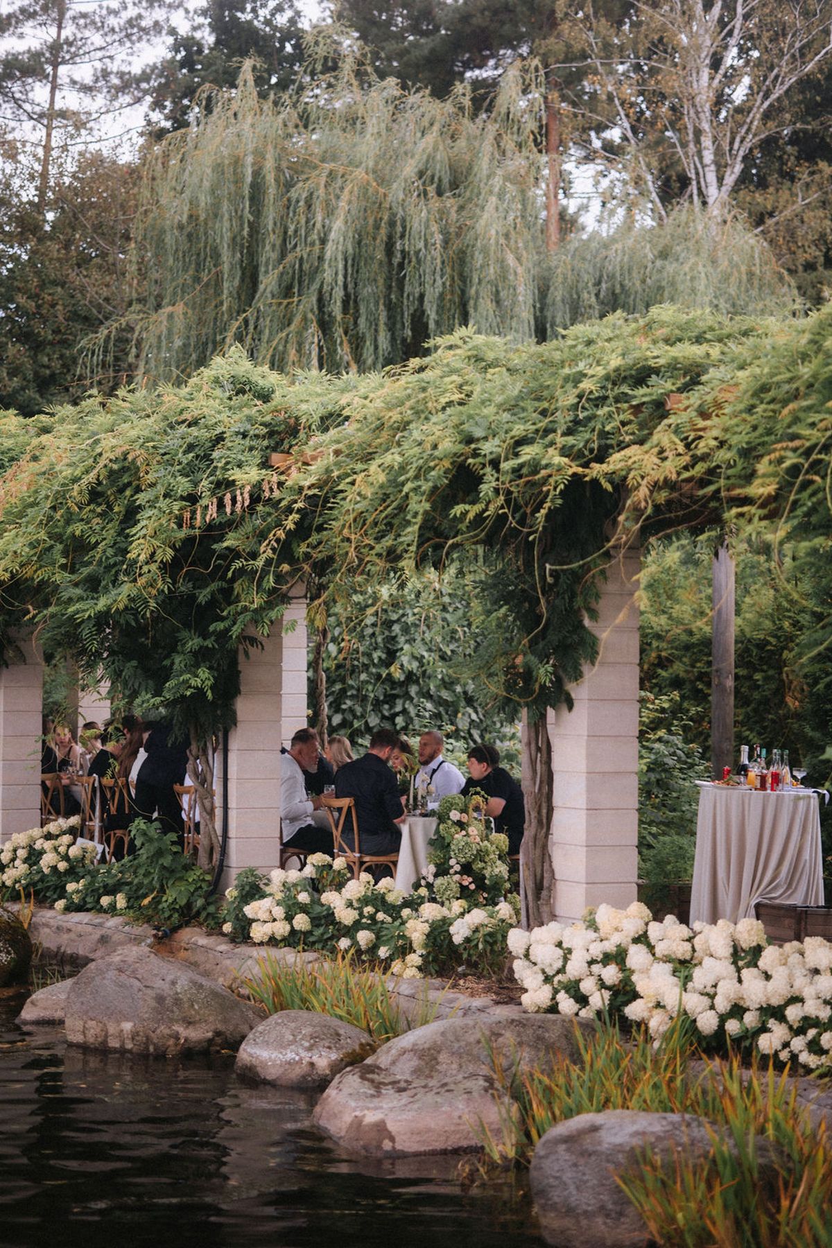 A romantic garden wedding ceremony setup with weeping willows, lush greenery, and white flowers along a pond with large rocks.