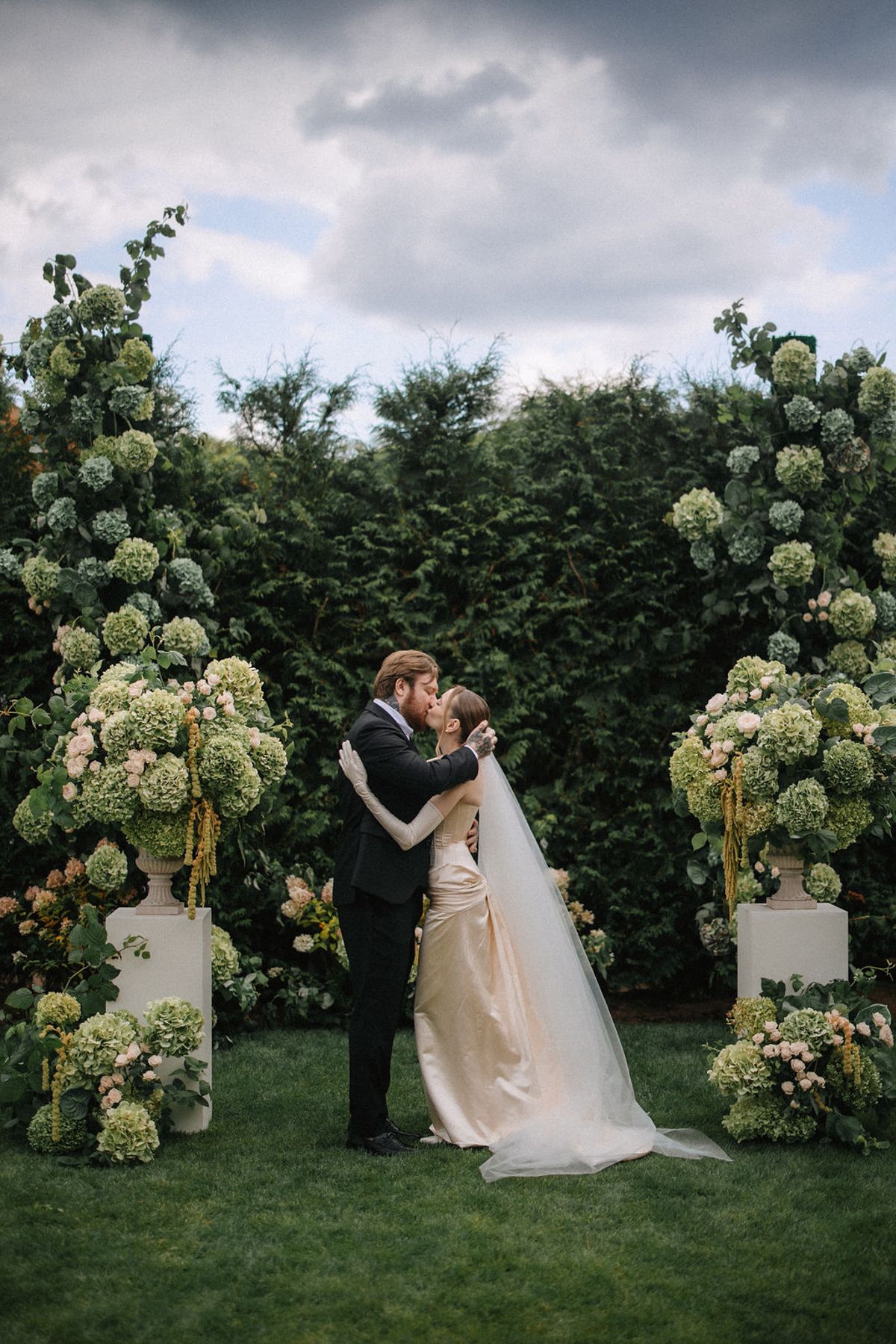 Elegant wedding portraits in a garden with white floral arrangements against a dramatic cloudy sky.