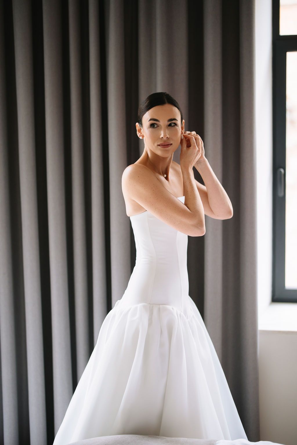 A graceful bride in a white ballgown twirls elegantly in front of floor-length curtains in a bridal suite.
