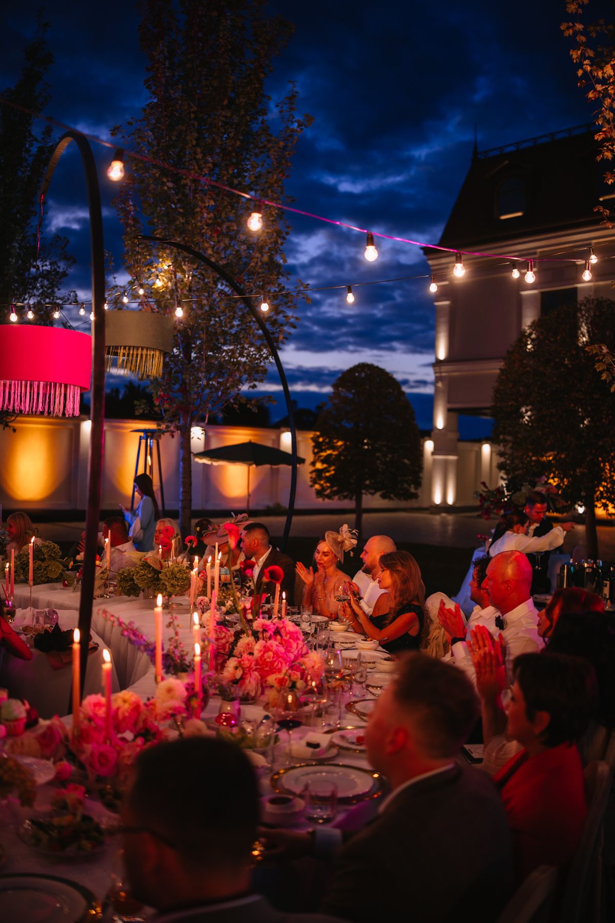 Outdoor evening wedding reception with string lights and pink floral centerpieces on tables under a dramatic twilight sky.