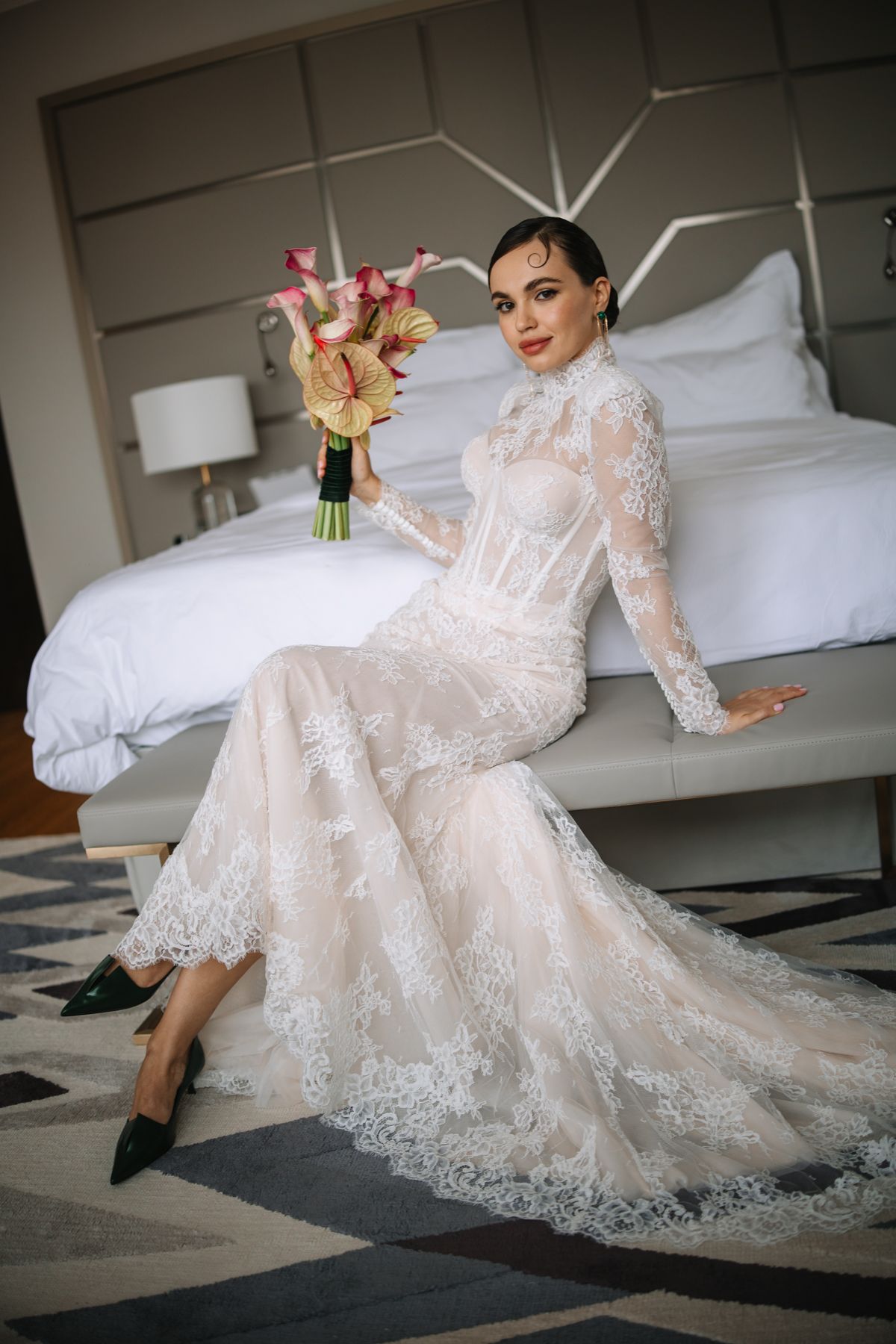 A bride in a flowing lace wedding gown with long sleeves sits elegantly on a floor in a light-filled room.