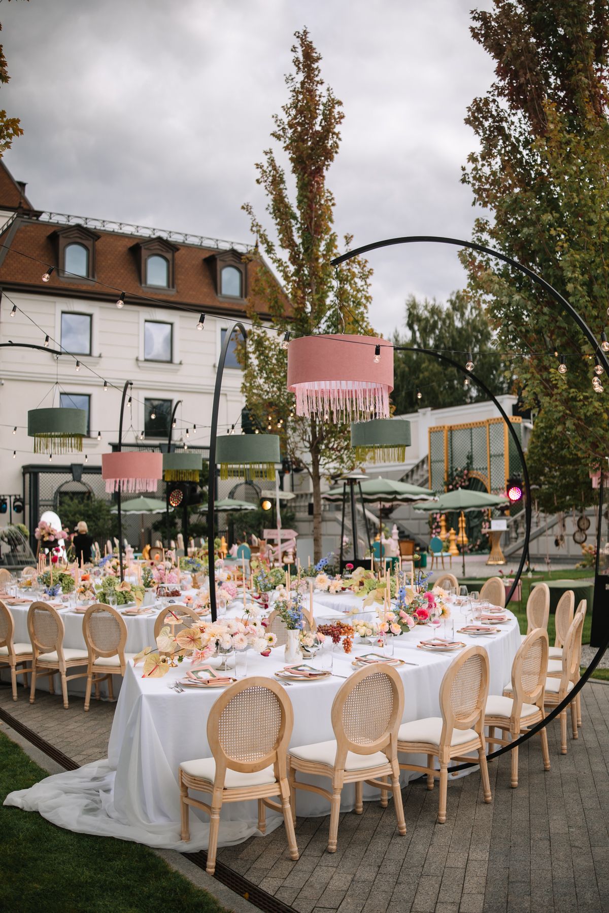 Elegant outdoor wedding reception setup with long tables, white linens, and cane-back chairs under tall trees at dusk.