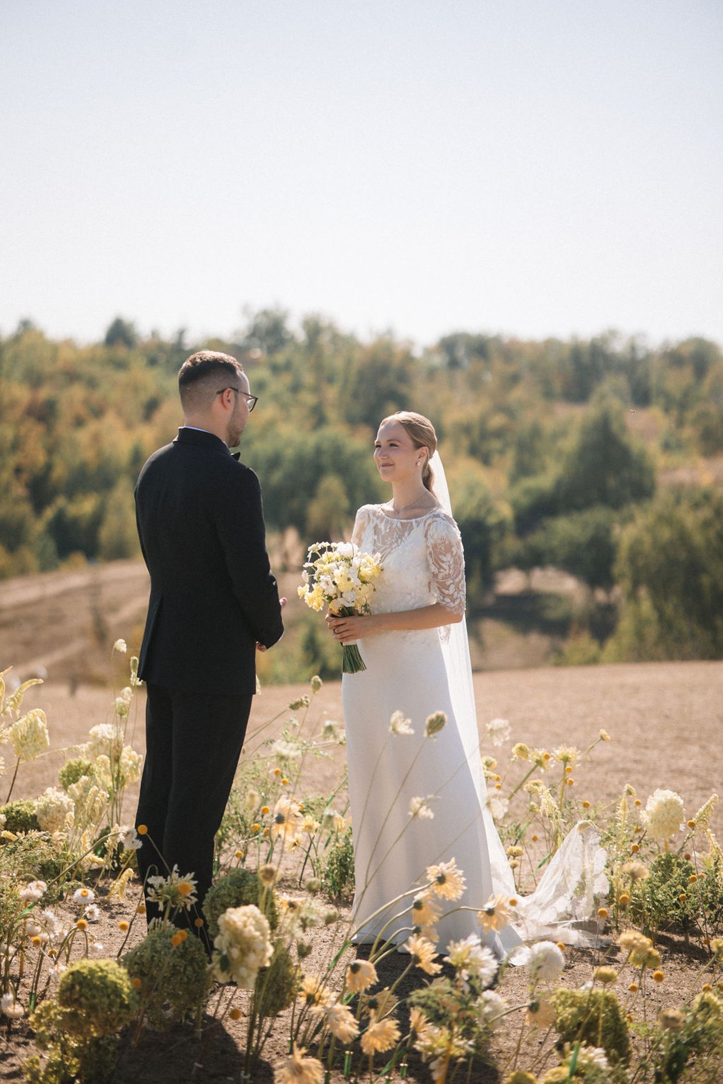 Newlywed couple standing among yellow wildflowers in a sunlit field during autumn.