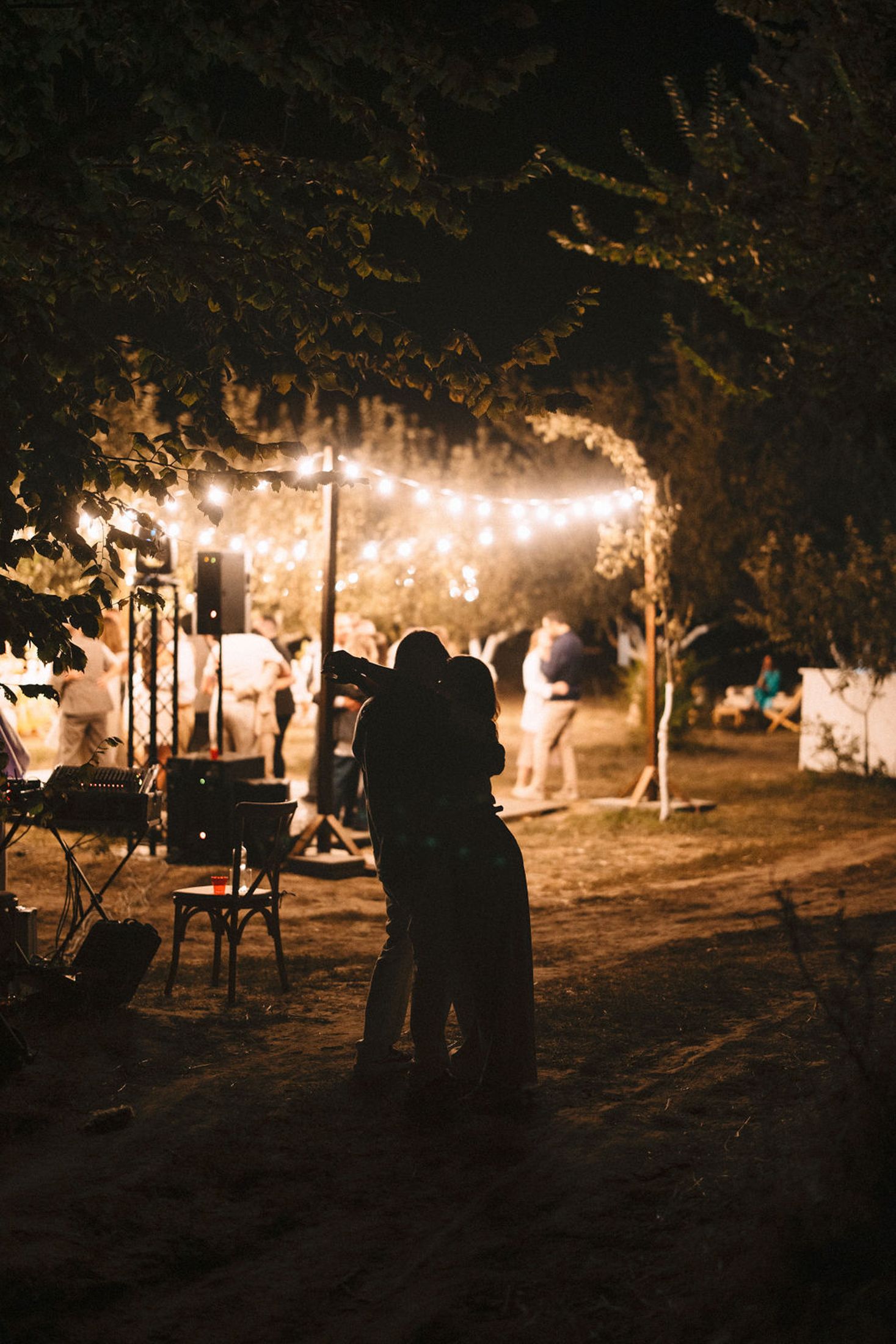 Silhouettes of people walking through an illuminated outdoor pathway with string lights hanging between trees at night.
