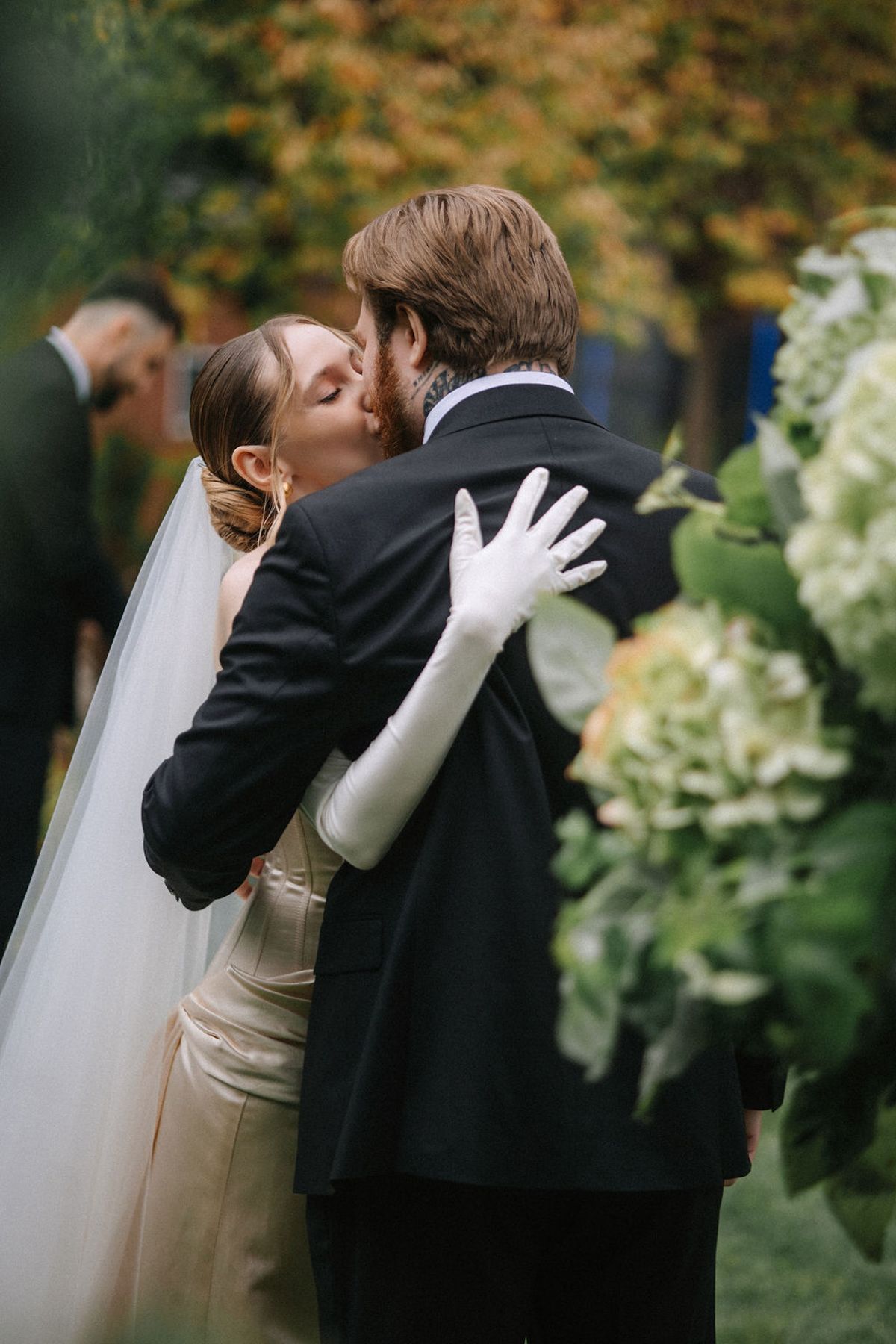 Romantic wedding couple shares intimate kisses in a garden setting with autumn foliage in the background.
