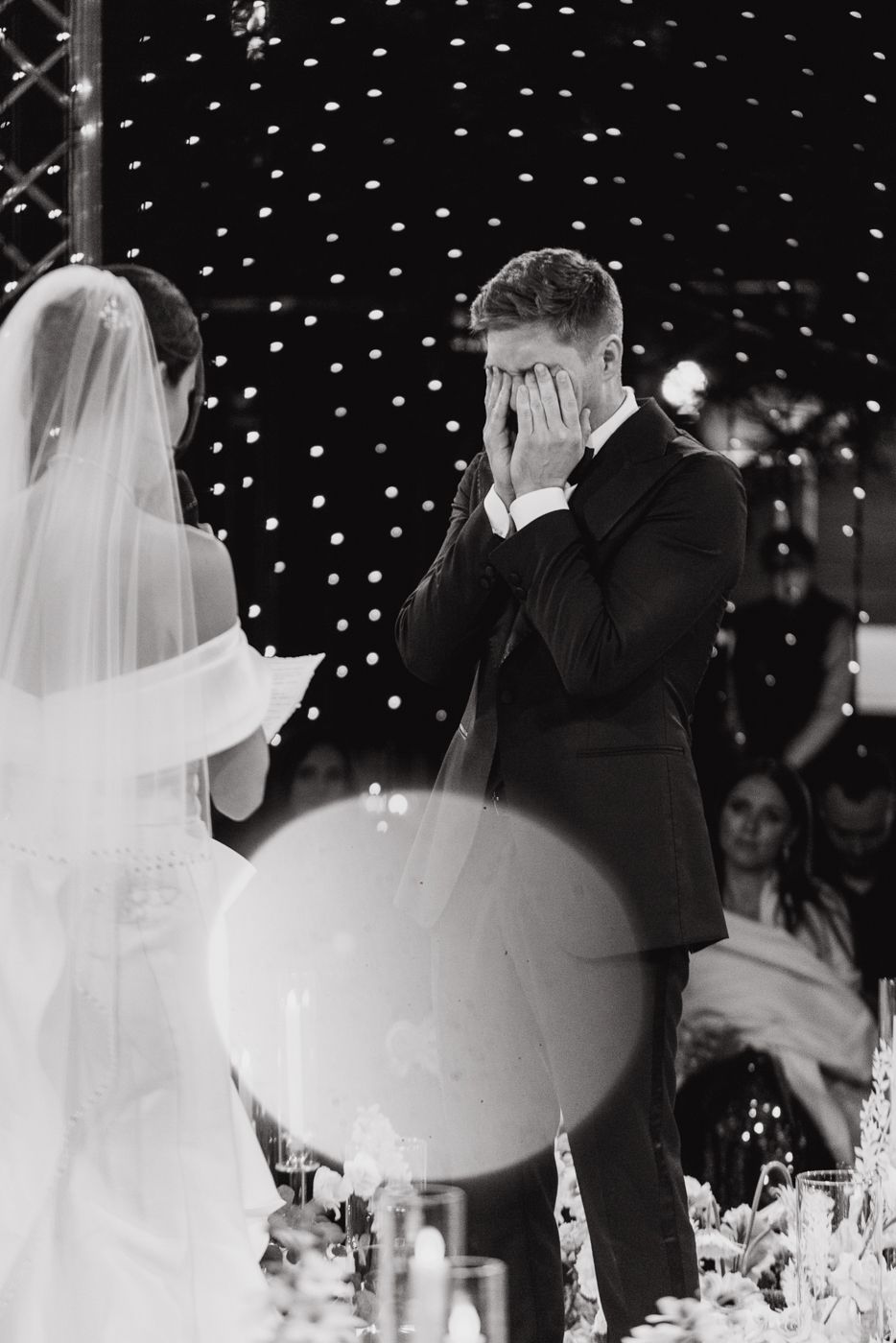 An emotional black and white moment during wedding vows with twinkling lights in the background.