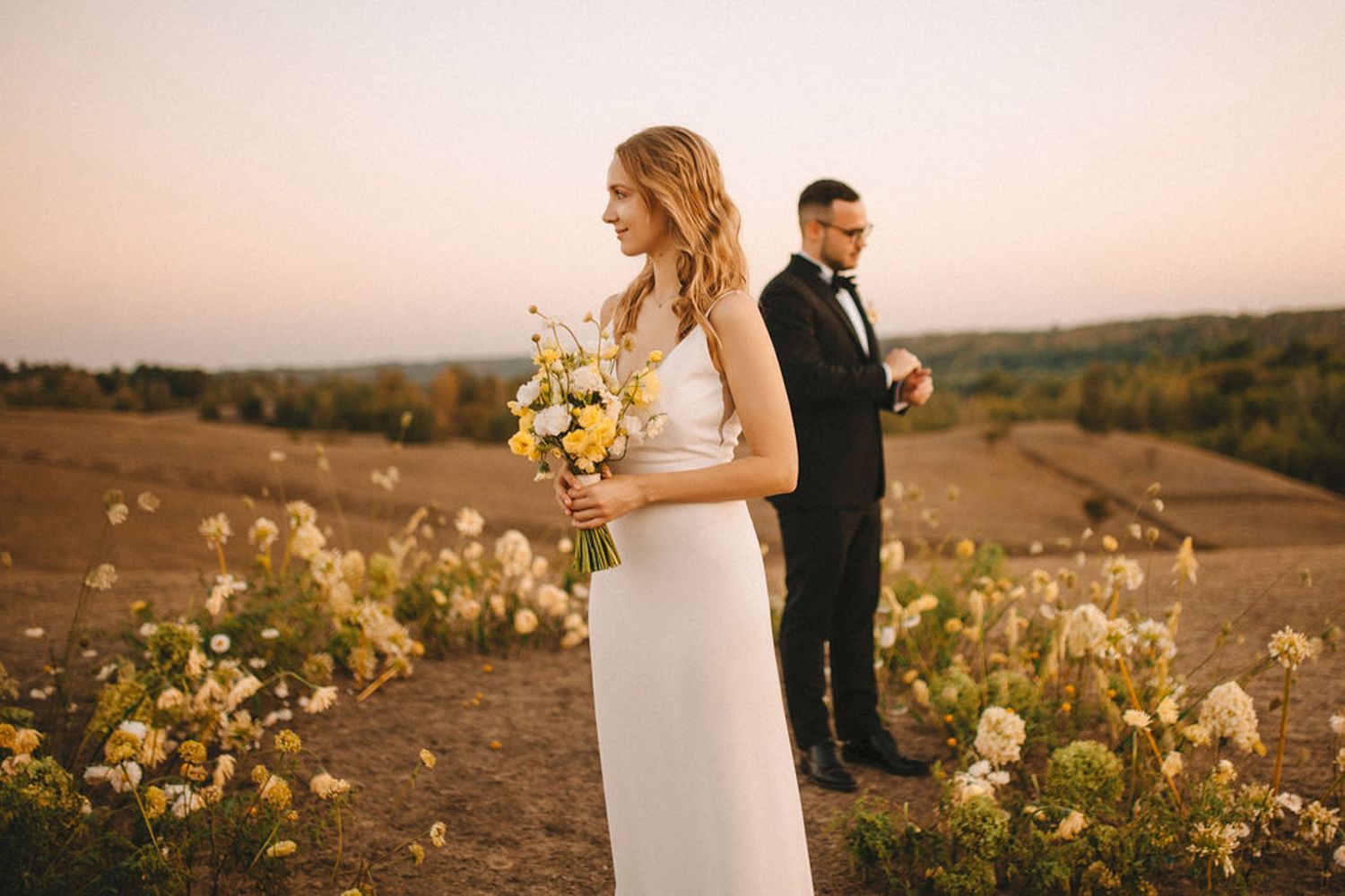 Newlywed couple walks hand in hand through golden vineyard at sunset with flowing white dress and formal attire.