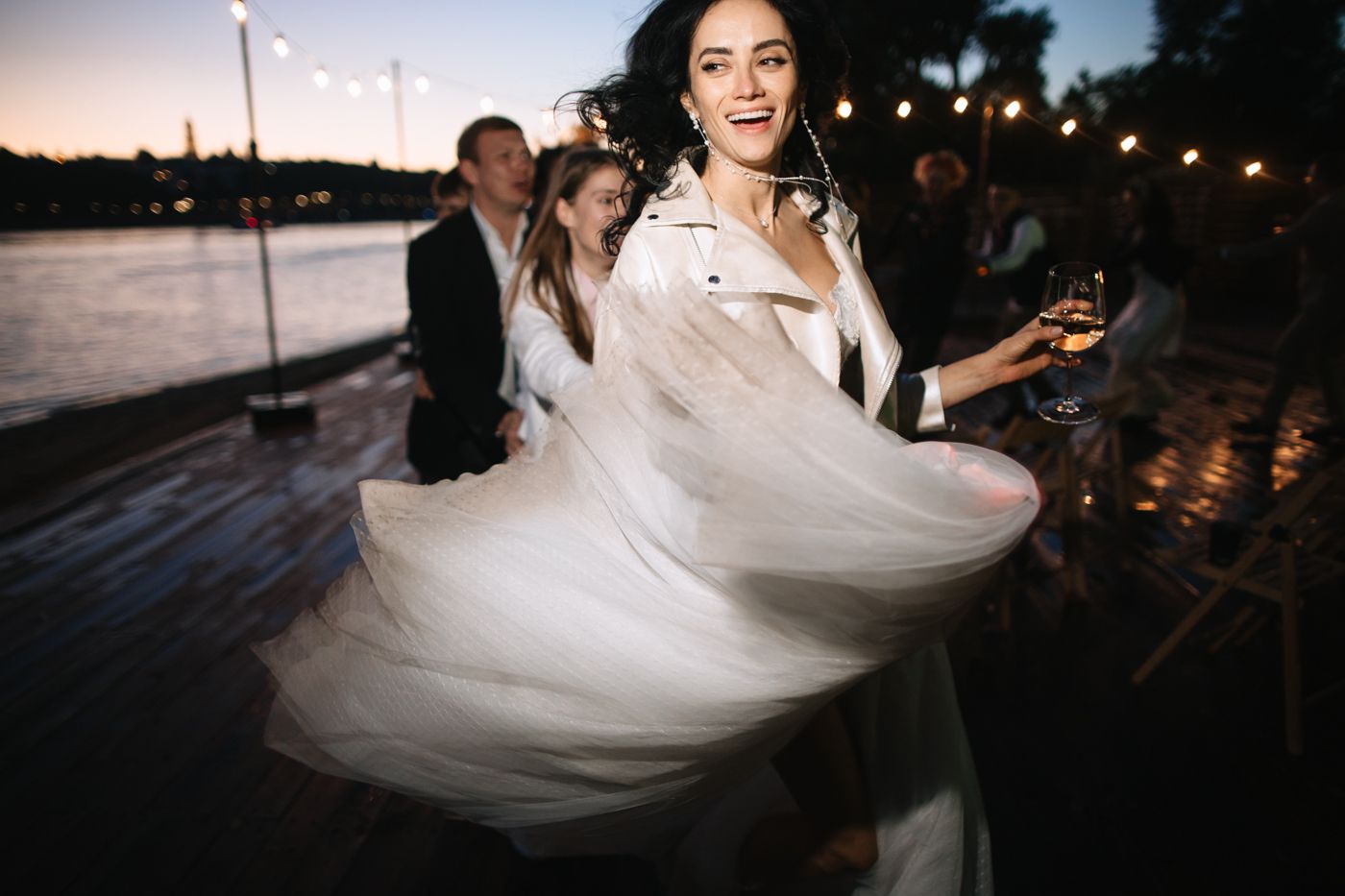 A bride in a flowing white dress twirls gracefully at dusk with string lights glowing in the background.