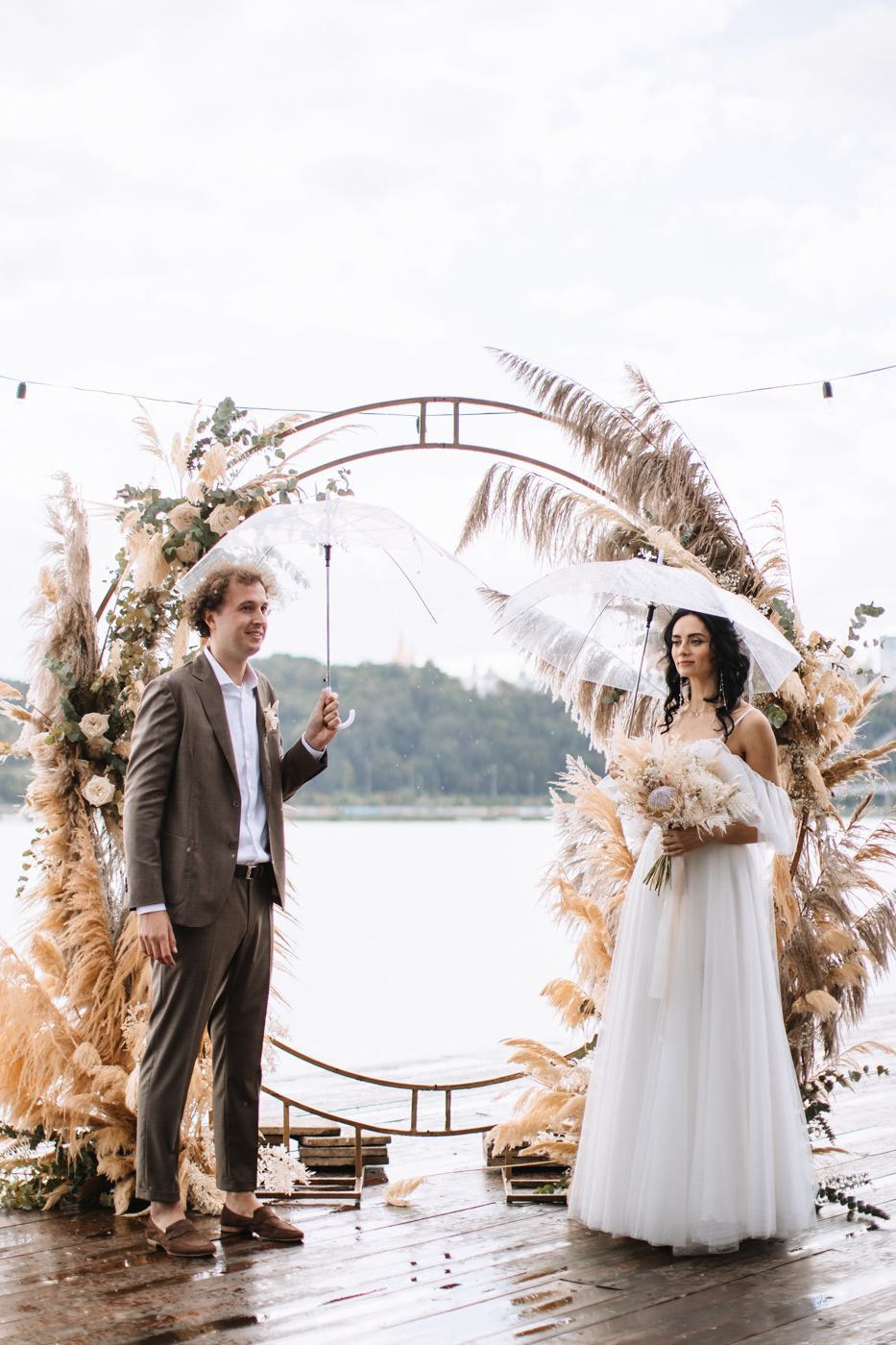 Wedding couple shares emotional first look moment on rainy dock decorated with pampas grass and floral arch.