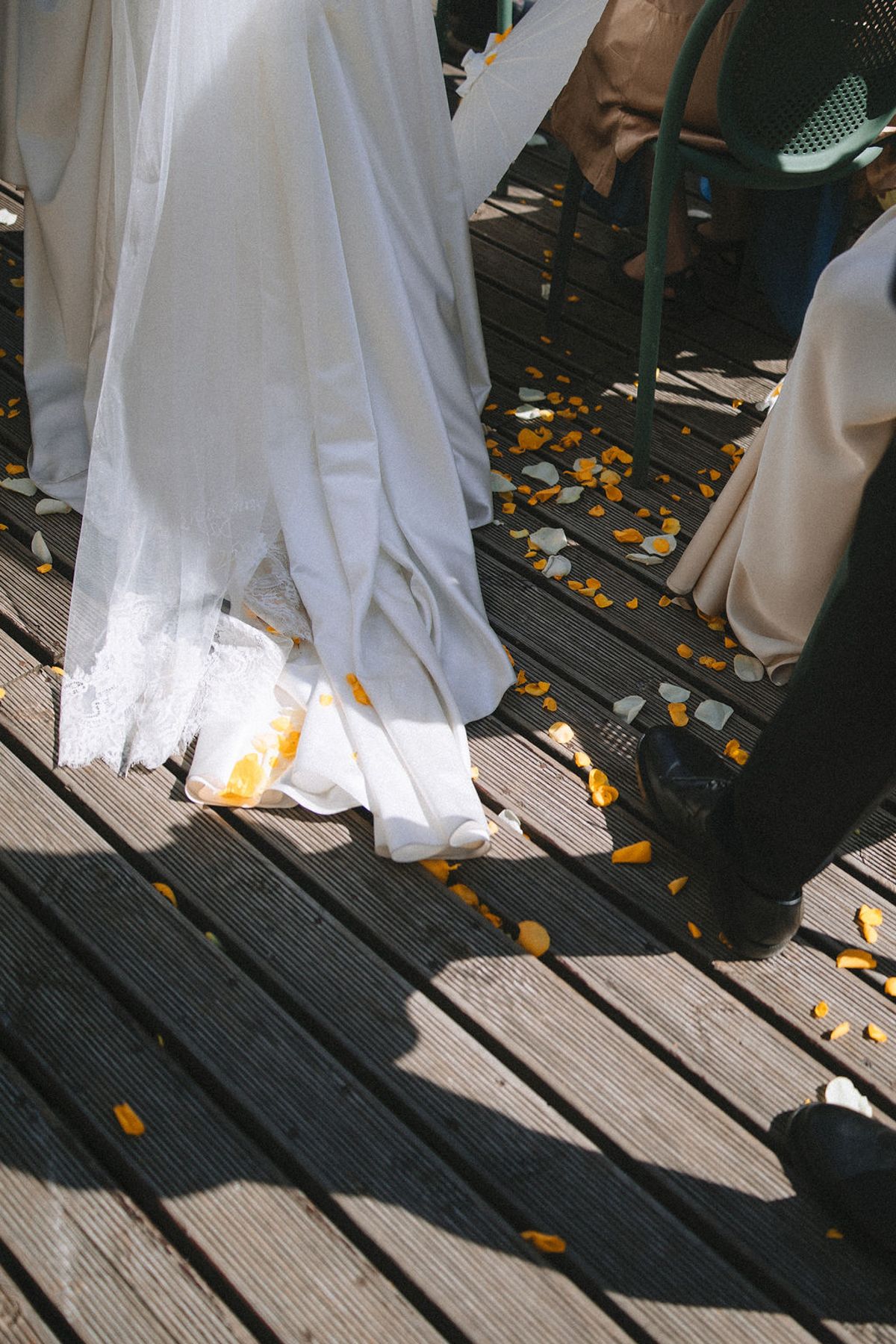 Orange flower petals scattered on wooden deck boards around flowing white wedding dress and dress shoes.