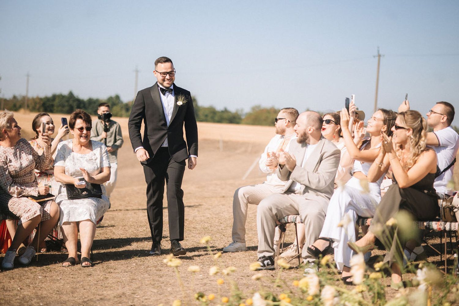 Wedding guests celebrate and toss flower petals during an outdoor ceremony in a rustic countryside setting.