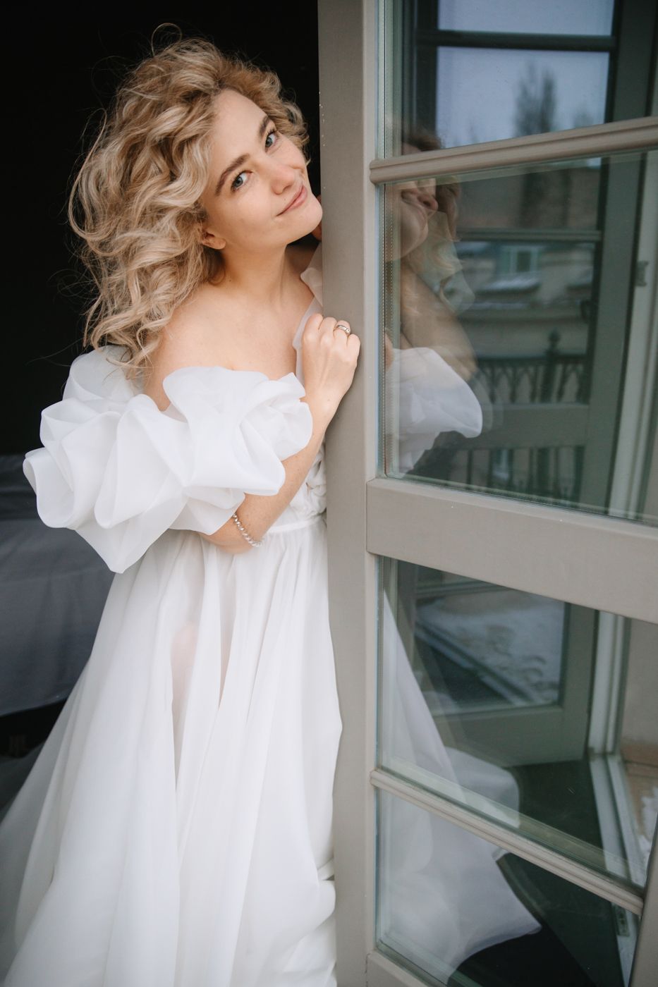 Woman in white flowing dress poses elegantly by window in photo series.