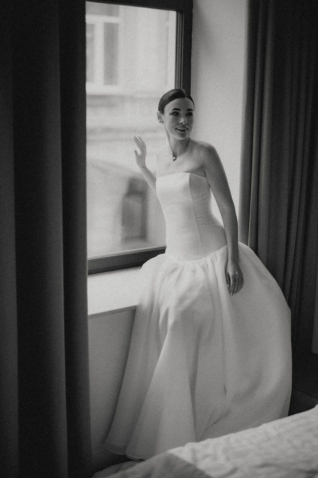 Black and white portrait of someone in a strapless wedding gown standing by a window.