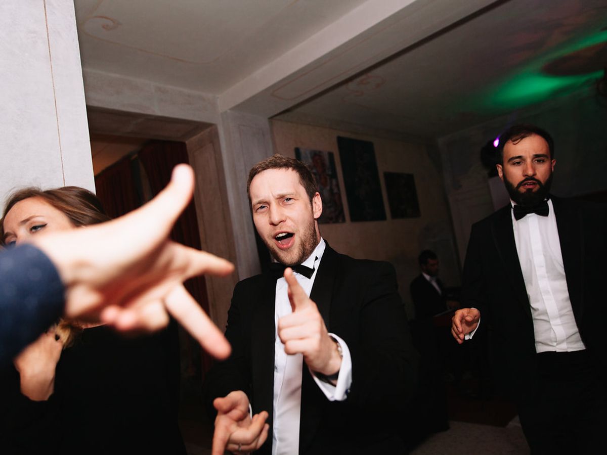 Wedding guests dance energetically in a dark room with white ceiling drapes and green lighting accents.