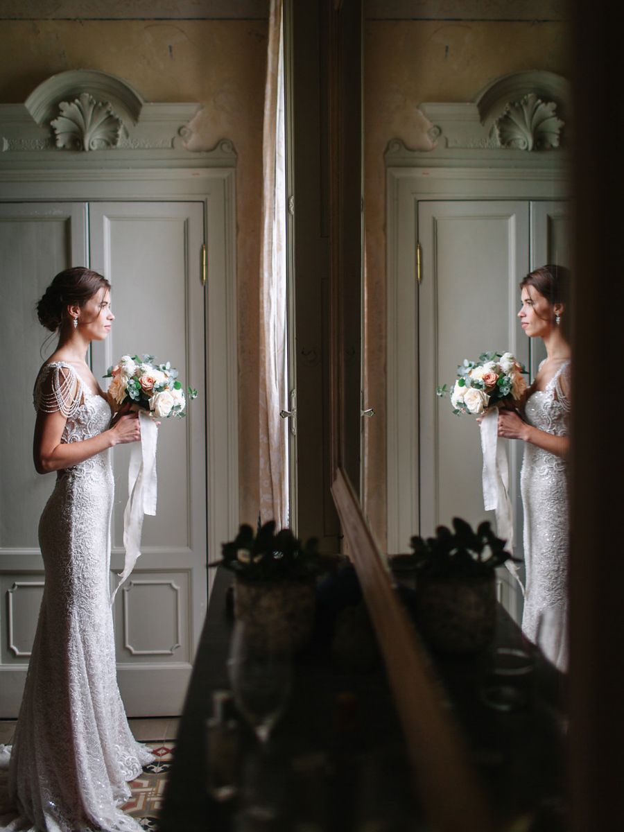 A bride in a white lace dress stands elegantly by a sunlit window holding a delicate floral bouquet.