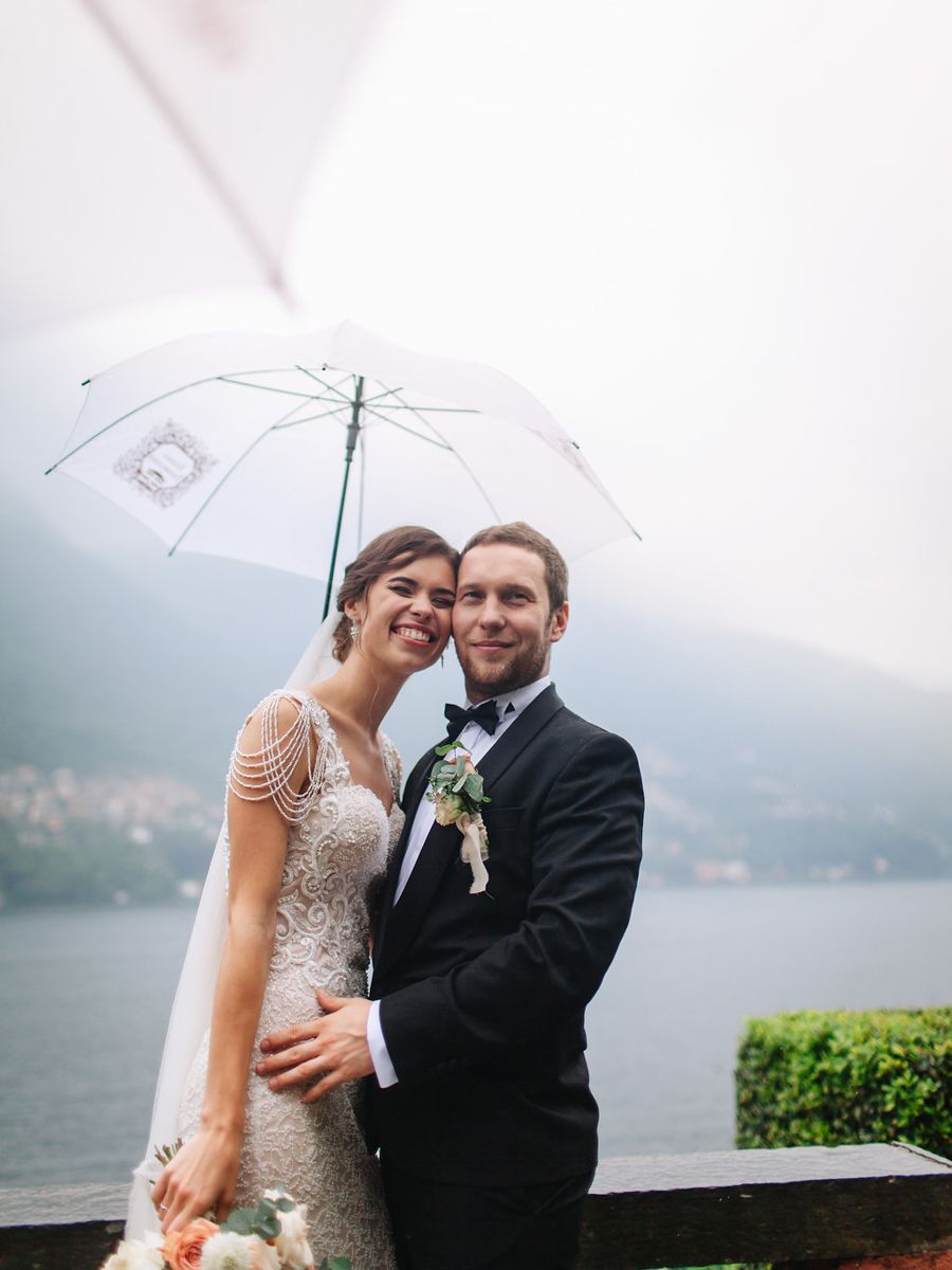 A couple shares an intimate moment under a white umbrella against a misty lakeside backdrop on their wedding day.