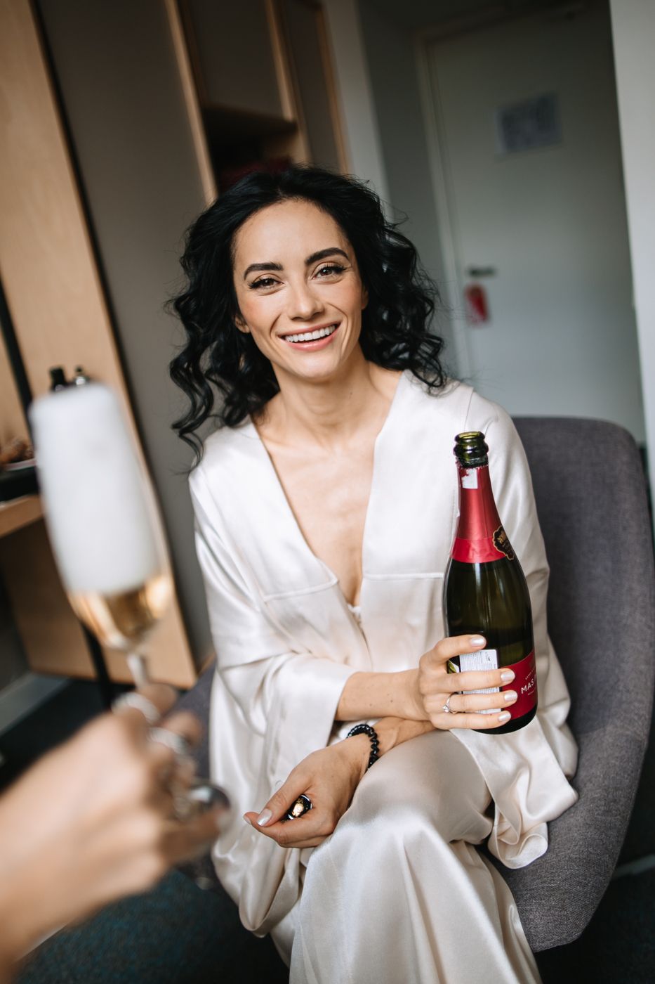 A person in a white silk robe holds a bottle while sitting and smiling during a candid photoshoot.