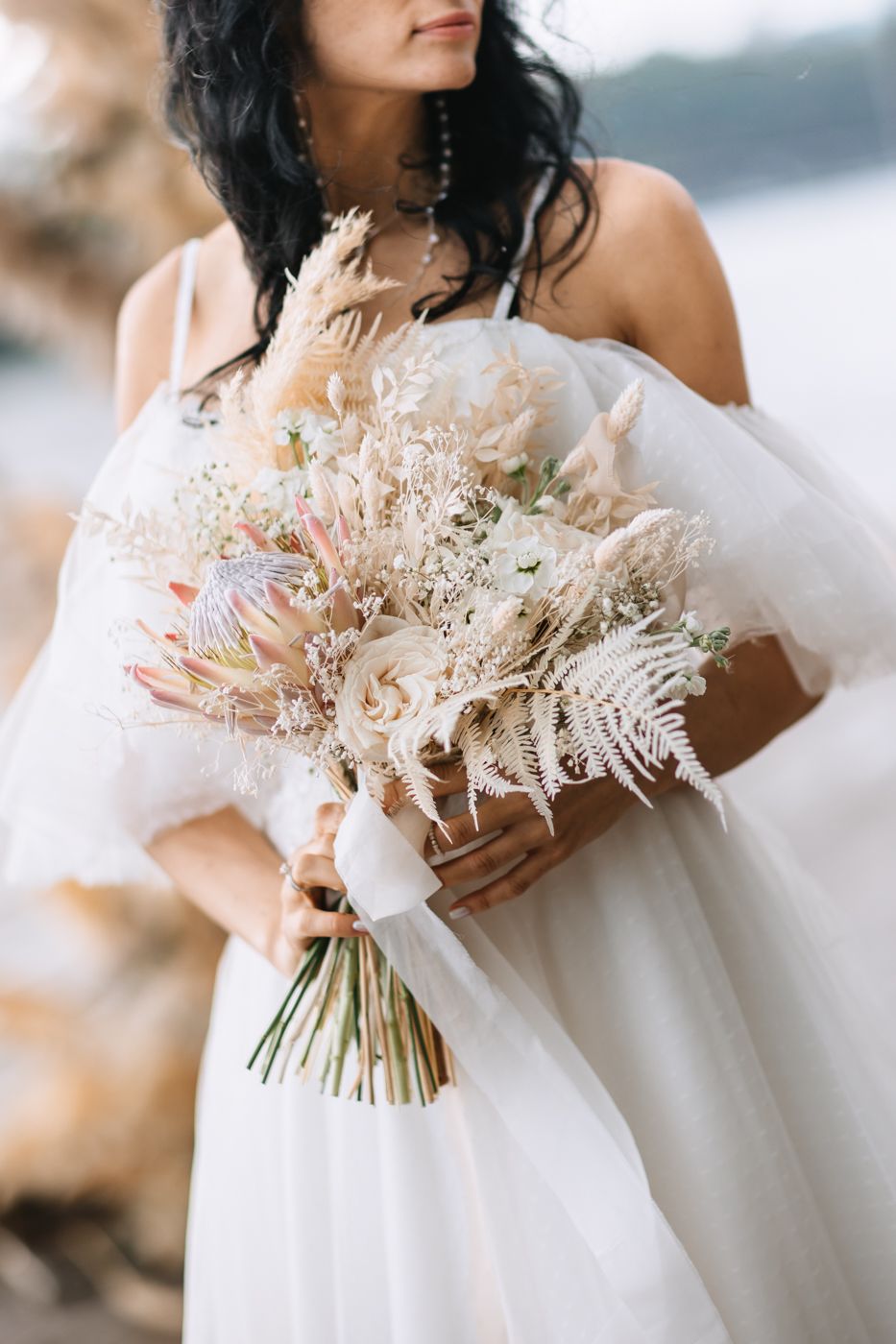 A delicate dried floral wedding bouquet with pampas grass and neutral-toned flowers held against a white dress.